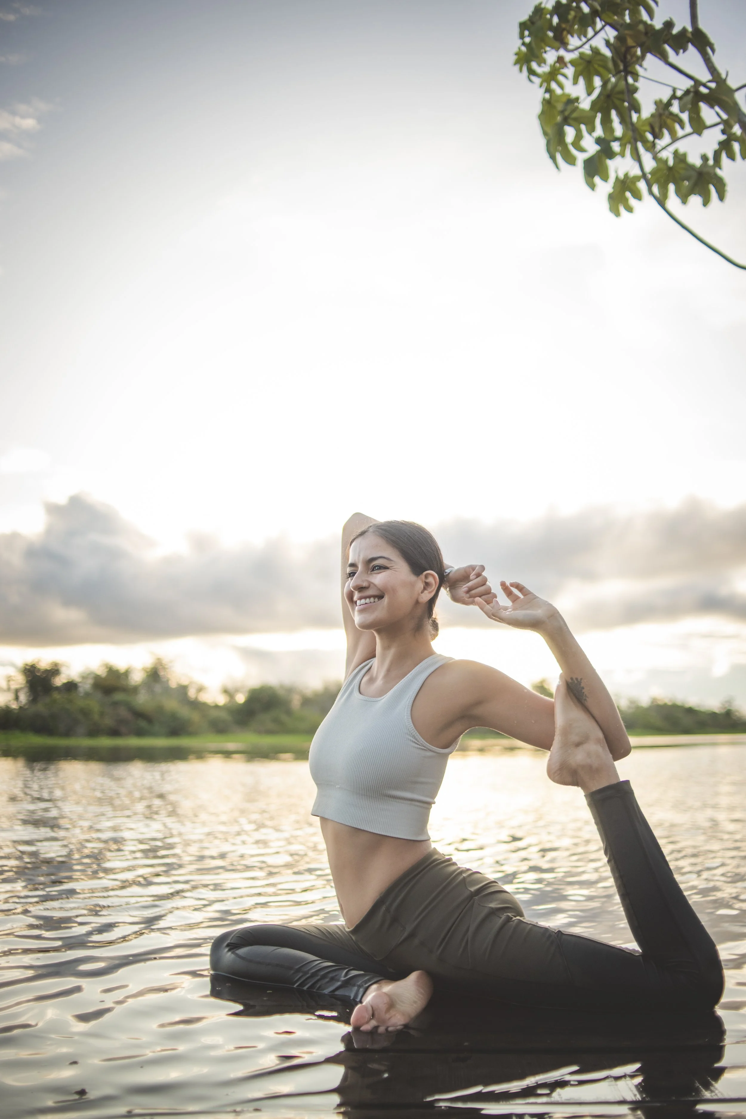 A woman practicing yoga on a floating platform in a lake during sunset, with a background of trees and cloudy sky.