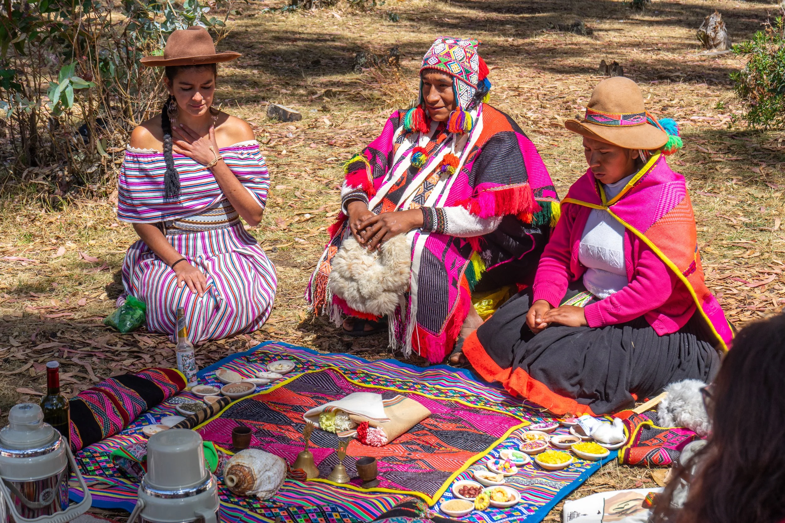 Three women dressed in colorful traditional clothing are sitting on a decorative blanket outdoors surrounded by various food items and drink bottles, engaging in a ceremony or ritual.