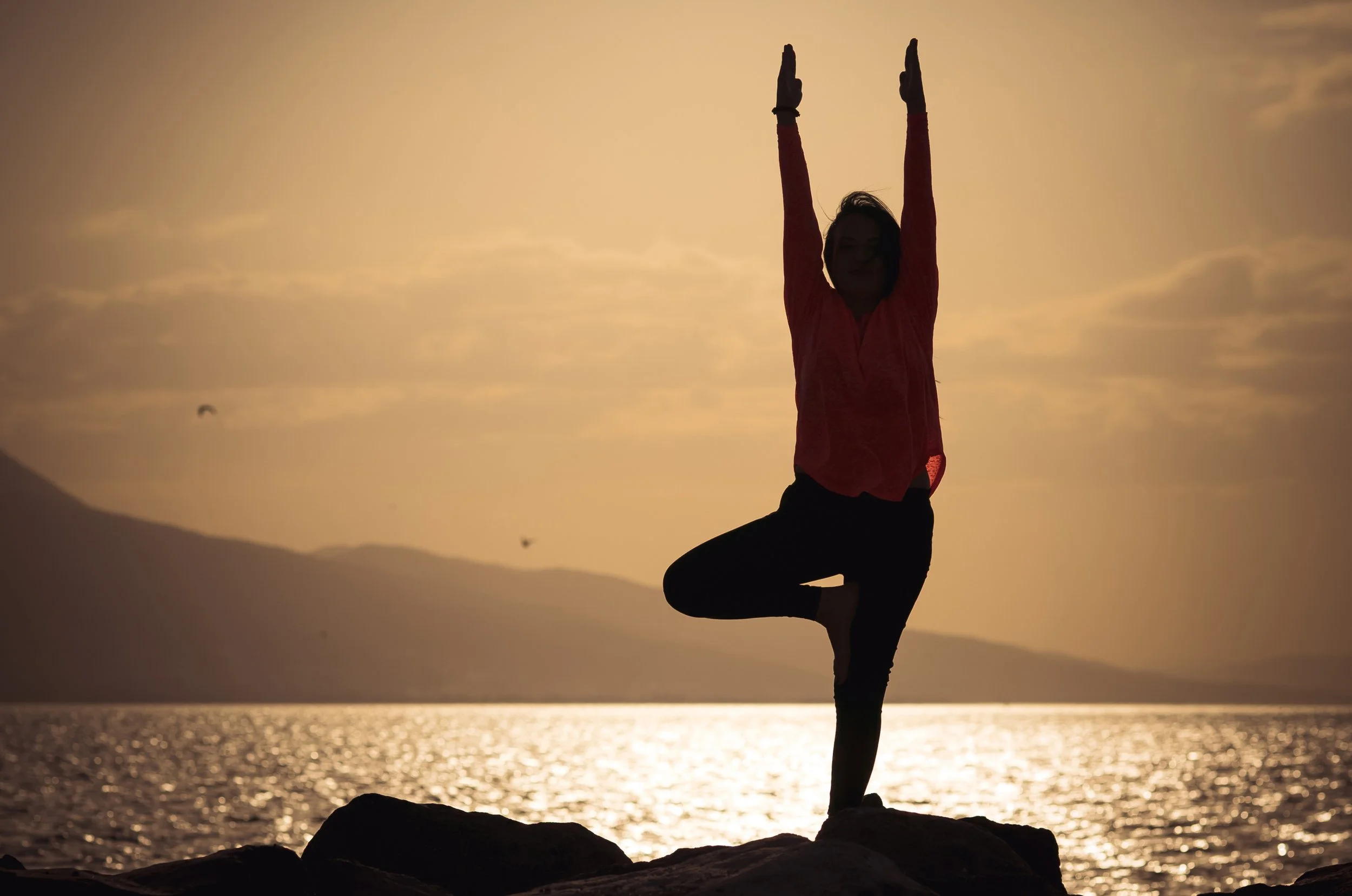 A person practicing yoga on one leg with hands raised above head at sunset on a beach.