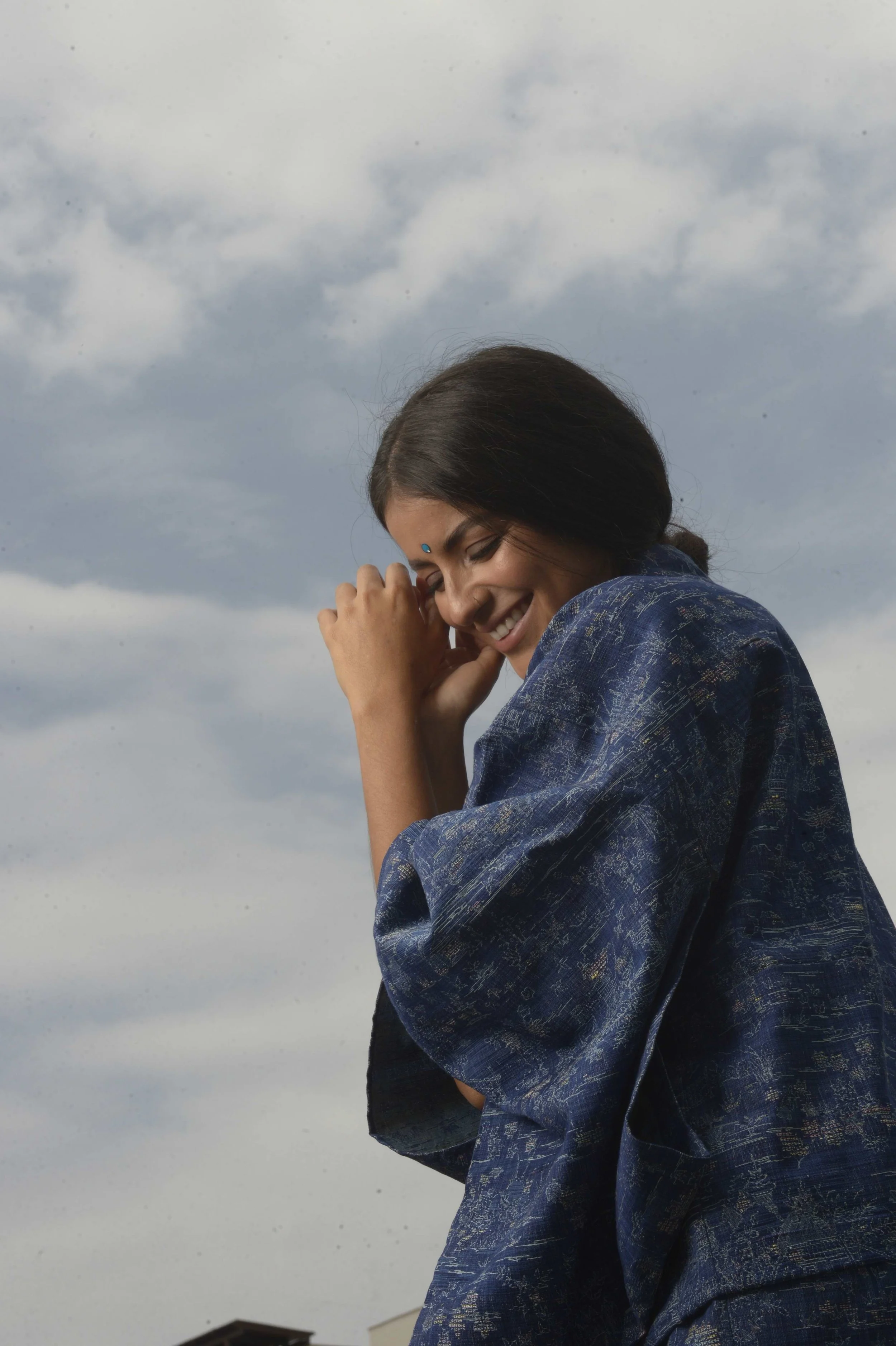 A woman with dark hair smiling and smiling with her hand near her face, wearing a blue traditional Indian sari, standing outdoors with cloudy sky in the background.