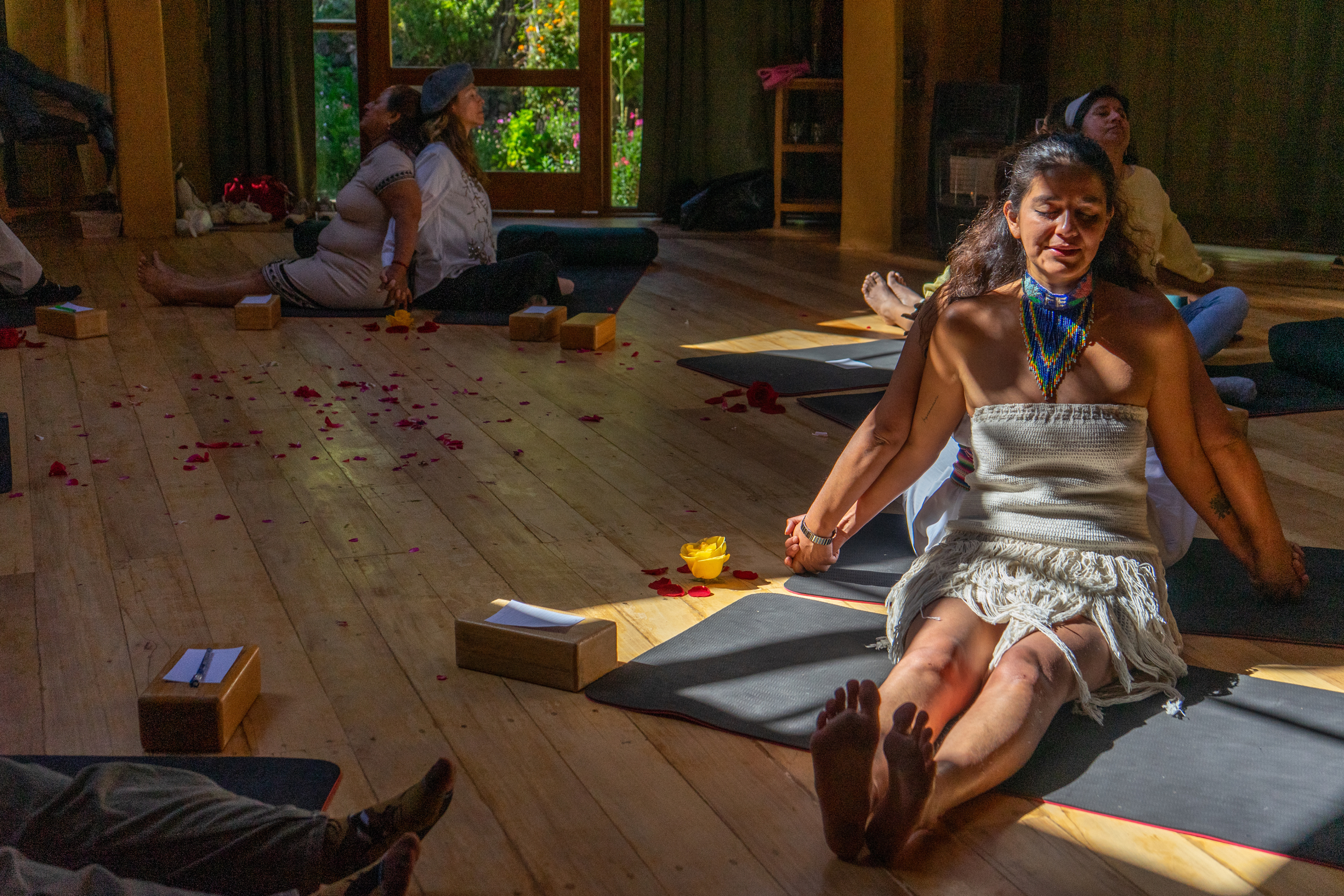 People engaging in a meditative or spiritual practice in a cozy room with wooden floors, yoga mats, and some decorative elements. They appear to be seated or lying in a relaxed, meditative pose with closed or focused eyes.