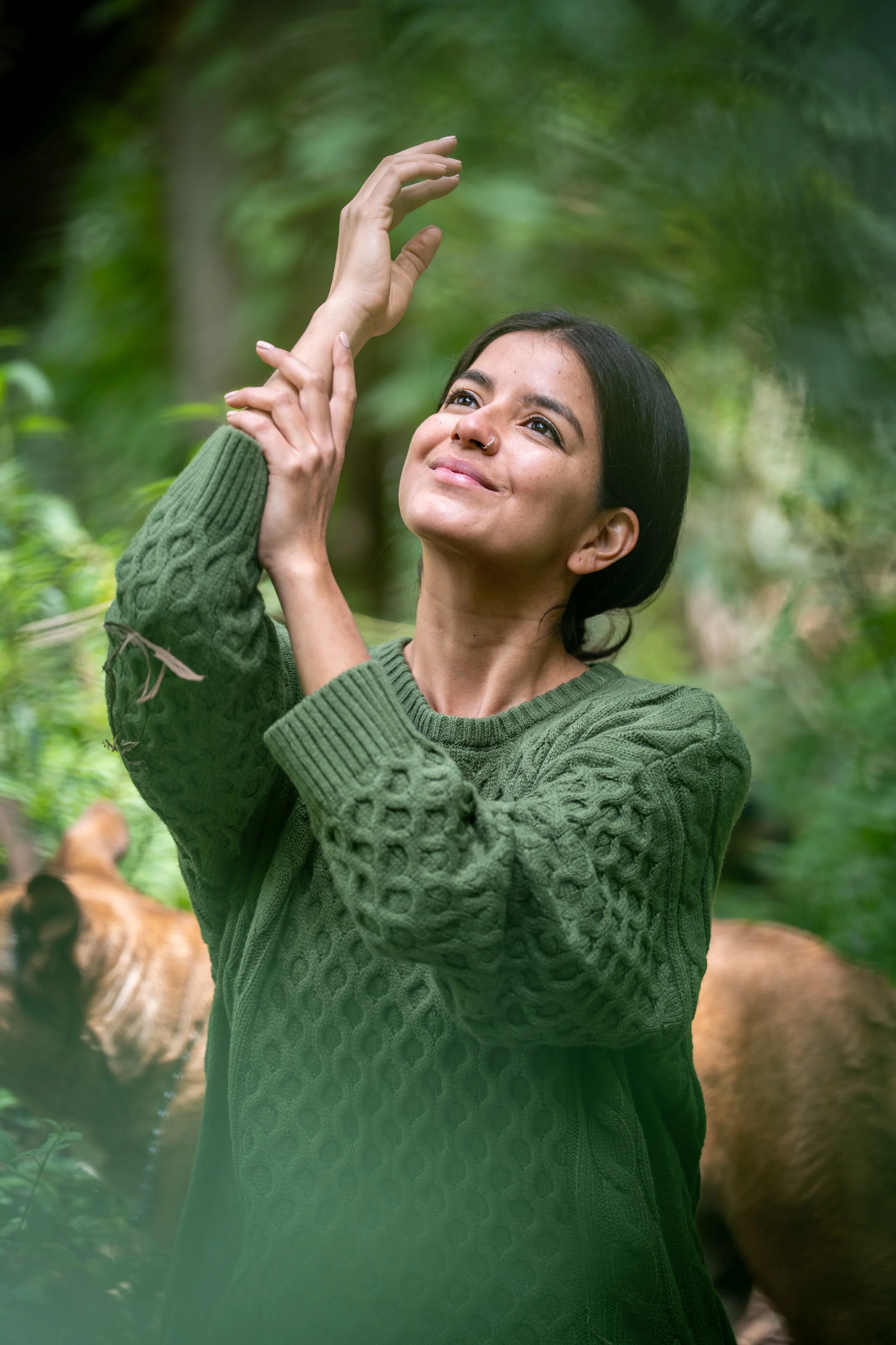 A woman with short black hair and a nose piercing standing outdoors in a green sweater, smiling and reaching up with her arms, with a blurred forest background and a tiger partially visible at the bottom of the frame.