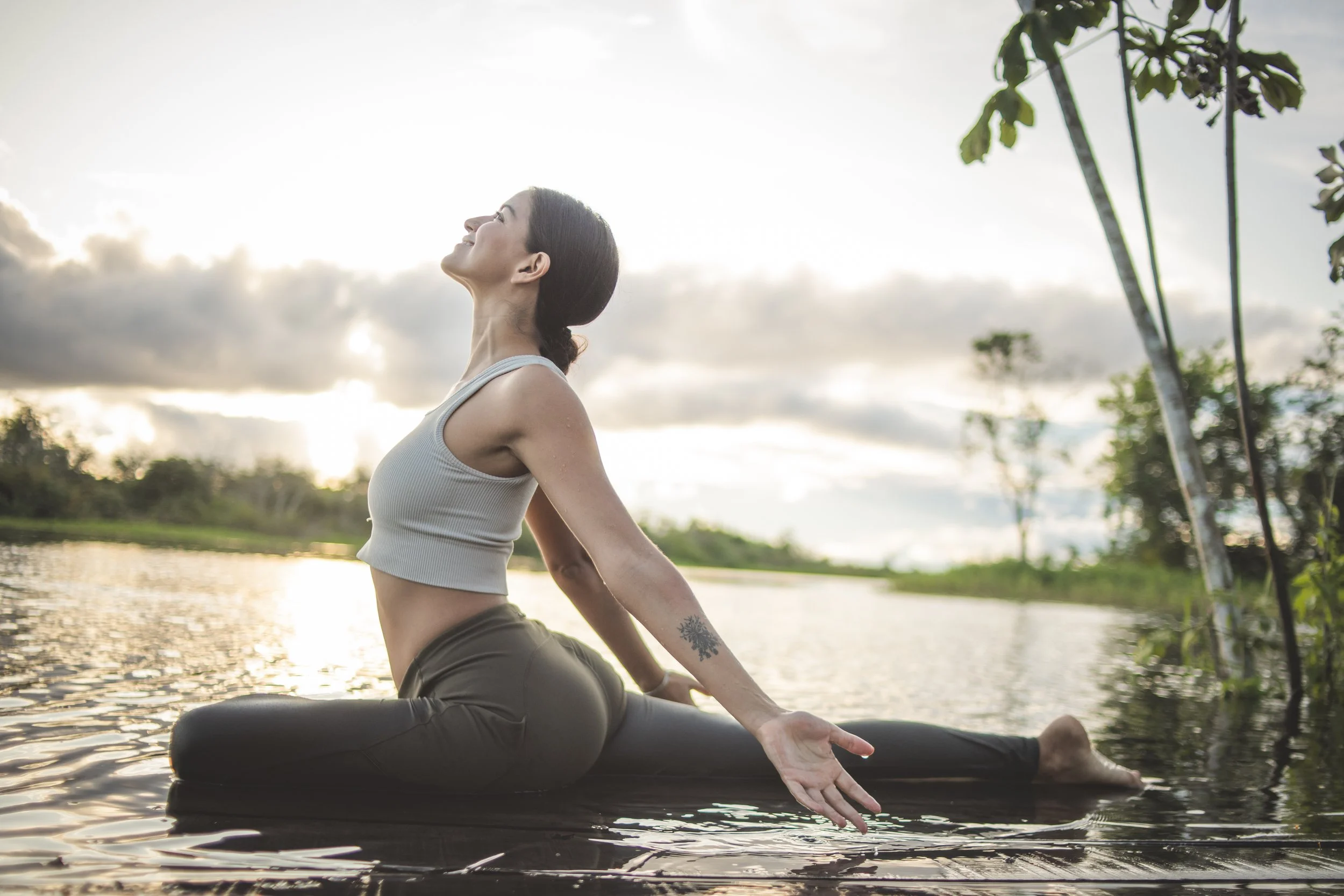 Woman practicing yoga outdoors on a mat near water during sunset, stretching with her eyes closed and smiling.