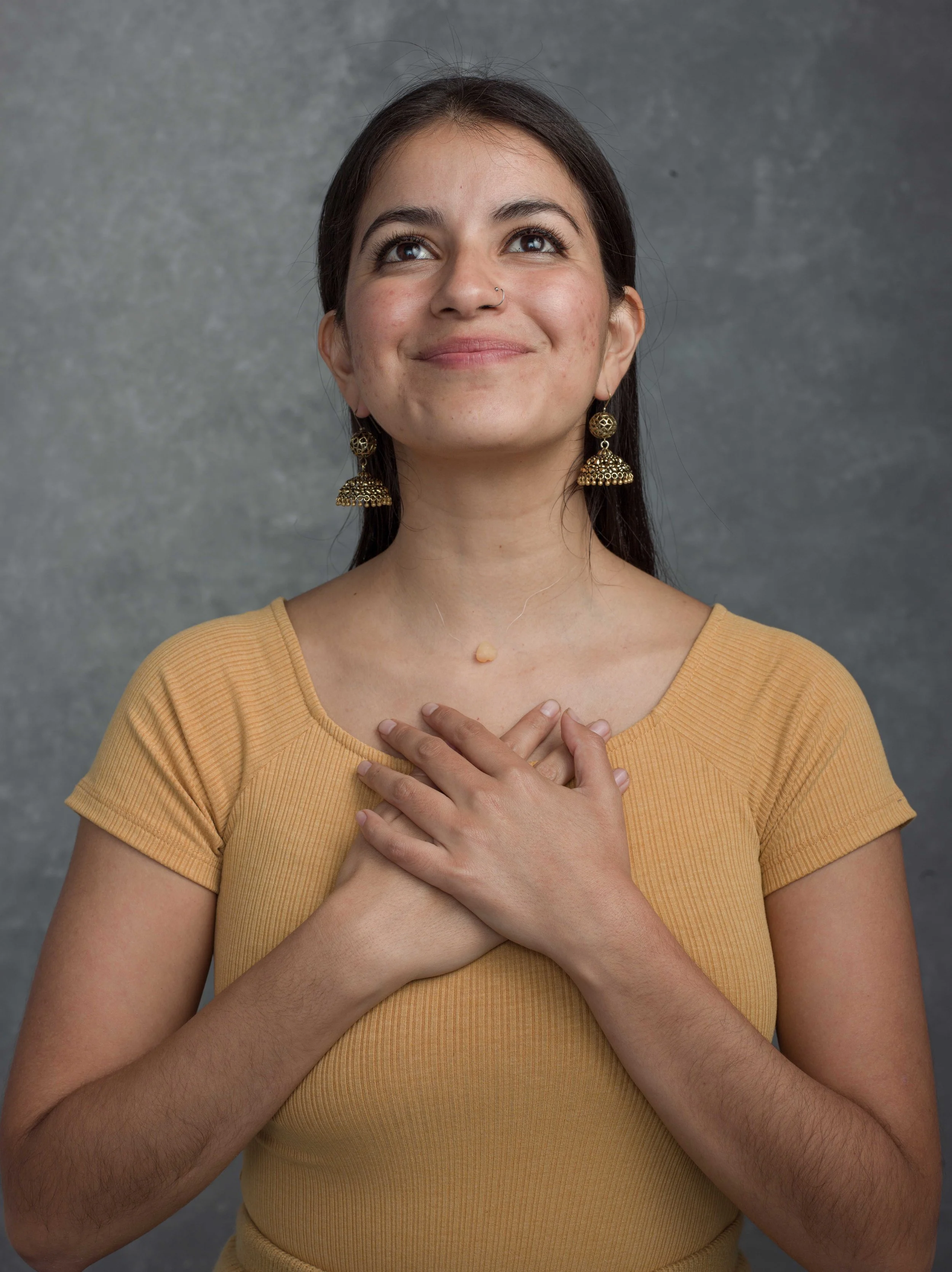 Young woman with dark hair, earrings, and a nose ring, wearing a yellow top, holding her hands over her chest, looking upward with a gentle smile against a gray background.