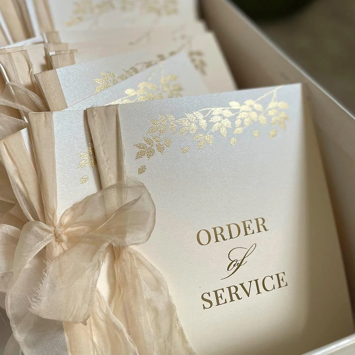 Elegant beige and gold service order envelopes with a sheer ribbon tied in a bow, adorned with gold leaf designs, arranged in a box.