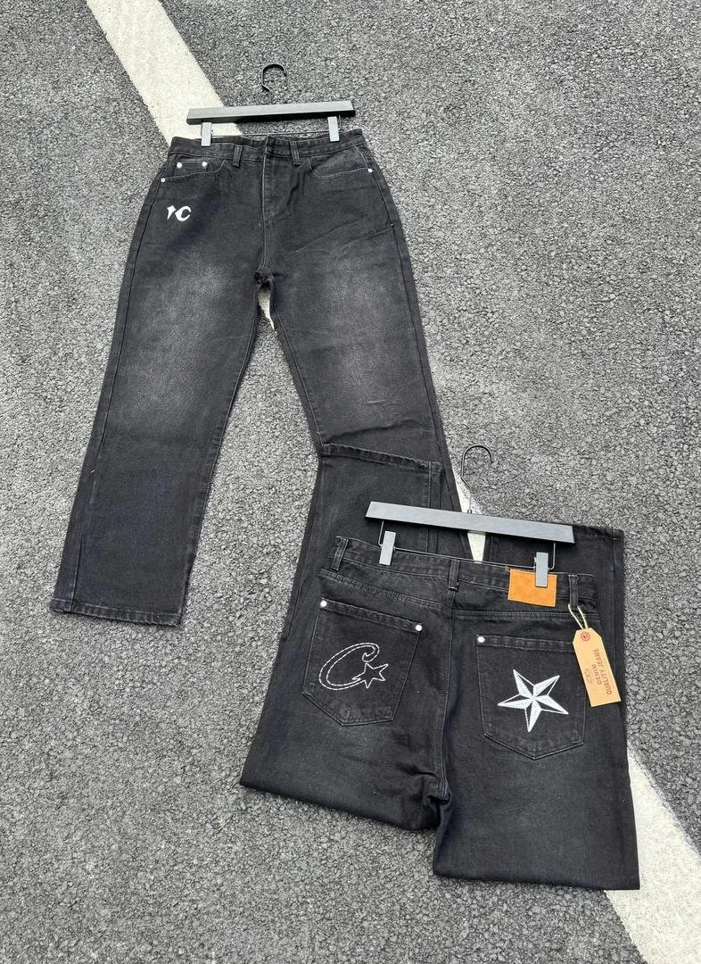 Two pairs of black jeans hanging on hangers on a gray textured surface, one pair facing front and the other facing back, showing embroidered designs.