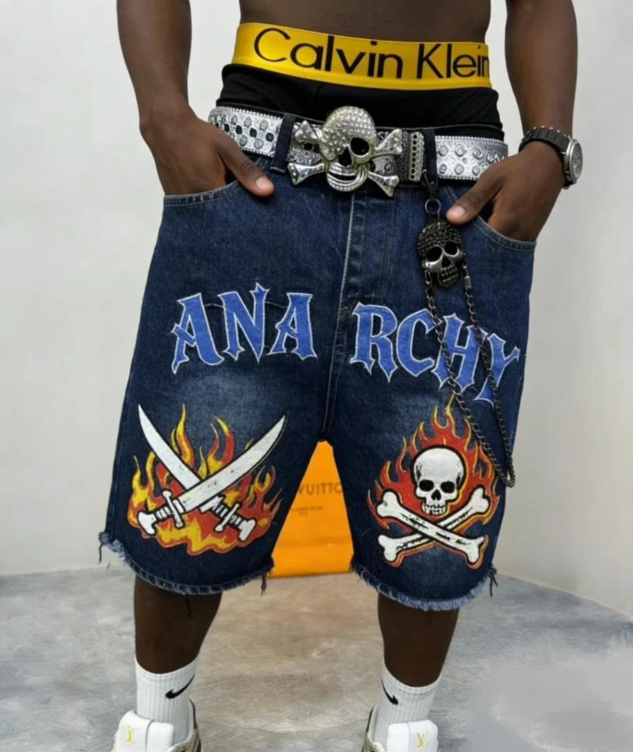 Person wearing dark blue denim shorts with white and blue embroidered text 'ANA RCHY' and flame and skull graphics, a yellow Calvin Klein waistband, a silver skull belt buckle, a skull chain accessory, white Nike socks, and white sneakers.