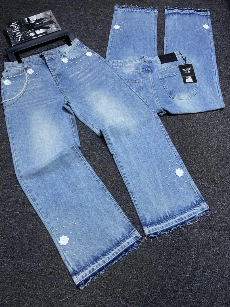 Two pairs of blue jeans with floral and pearl embellishments displayed on a dark surface, one pair with a black hanger and tag, the other folded.