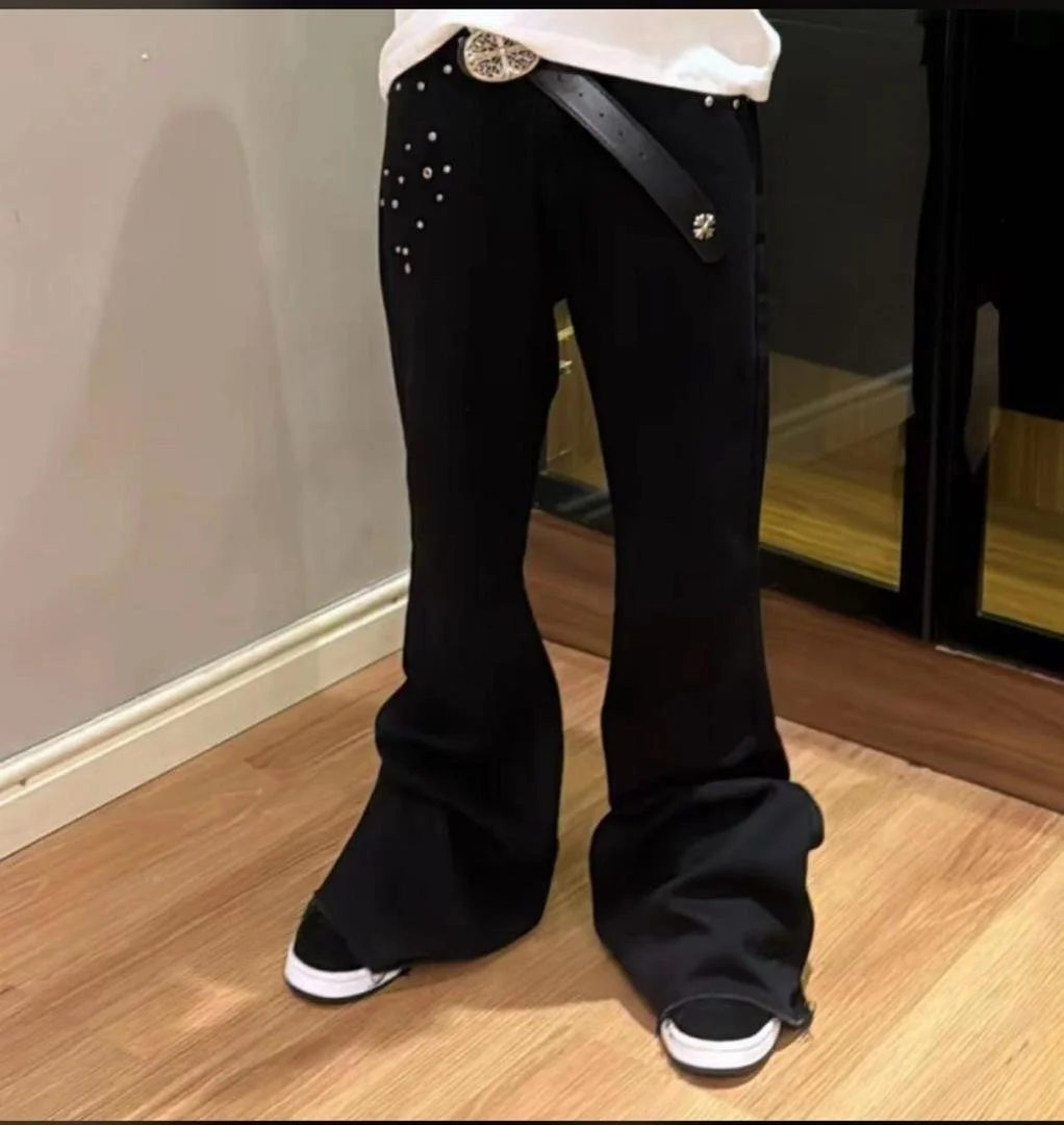 Person wearing black bell-bottom pants and black sneakers with white soles, standing on a wooden floor next to a mirror and a gray wall.