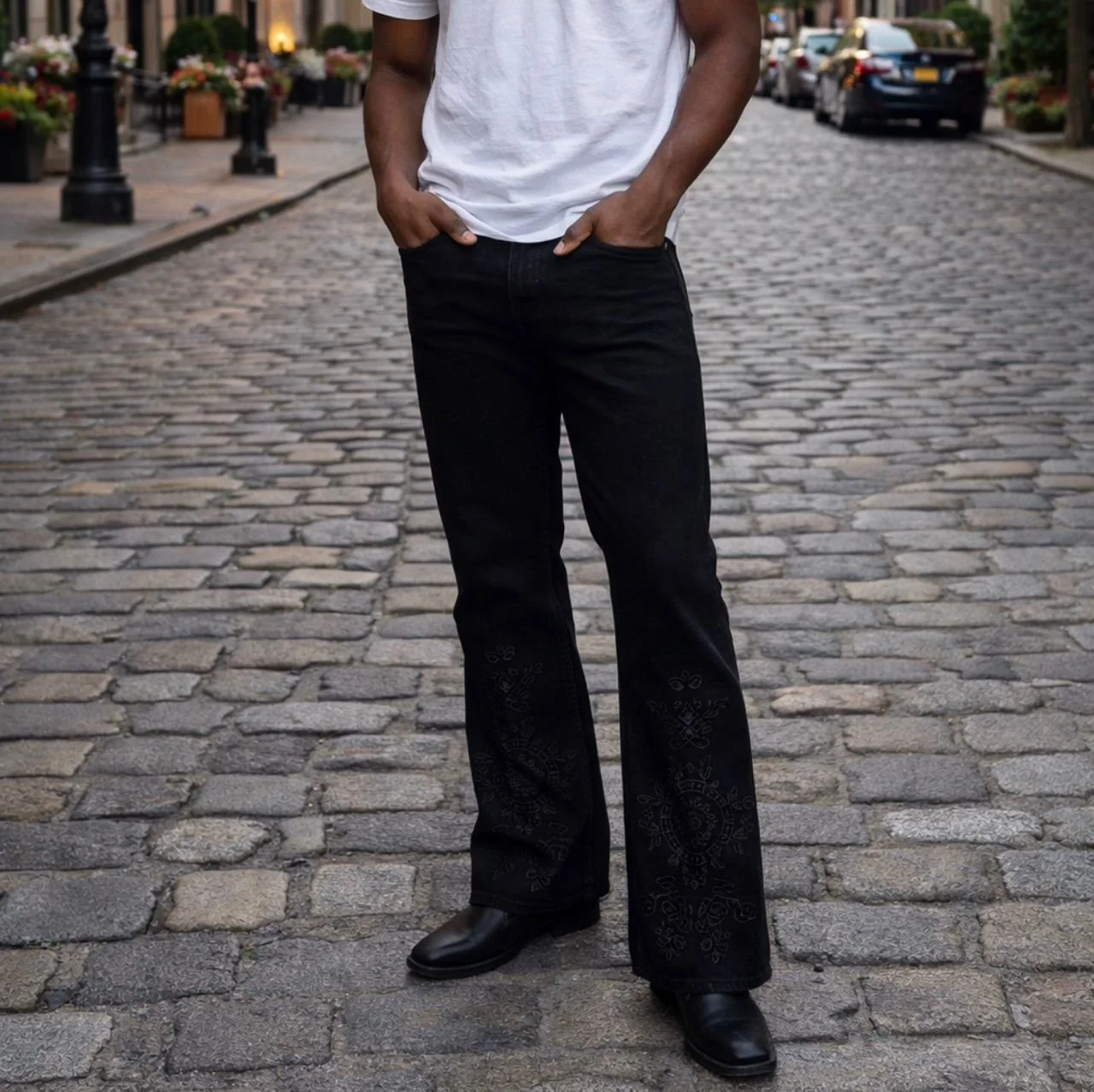 A person standing on a cobblestone street with hands in pockets, wearing a white T-shirt, black jeans with embroidered patterns, and black boots, with parked cars and flowerpots in the background.