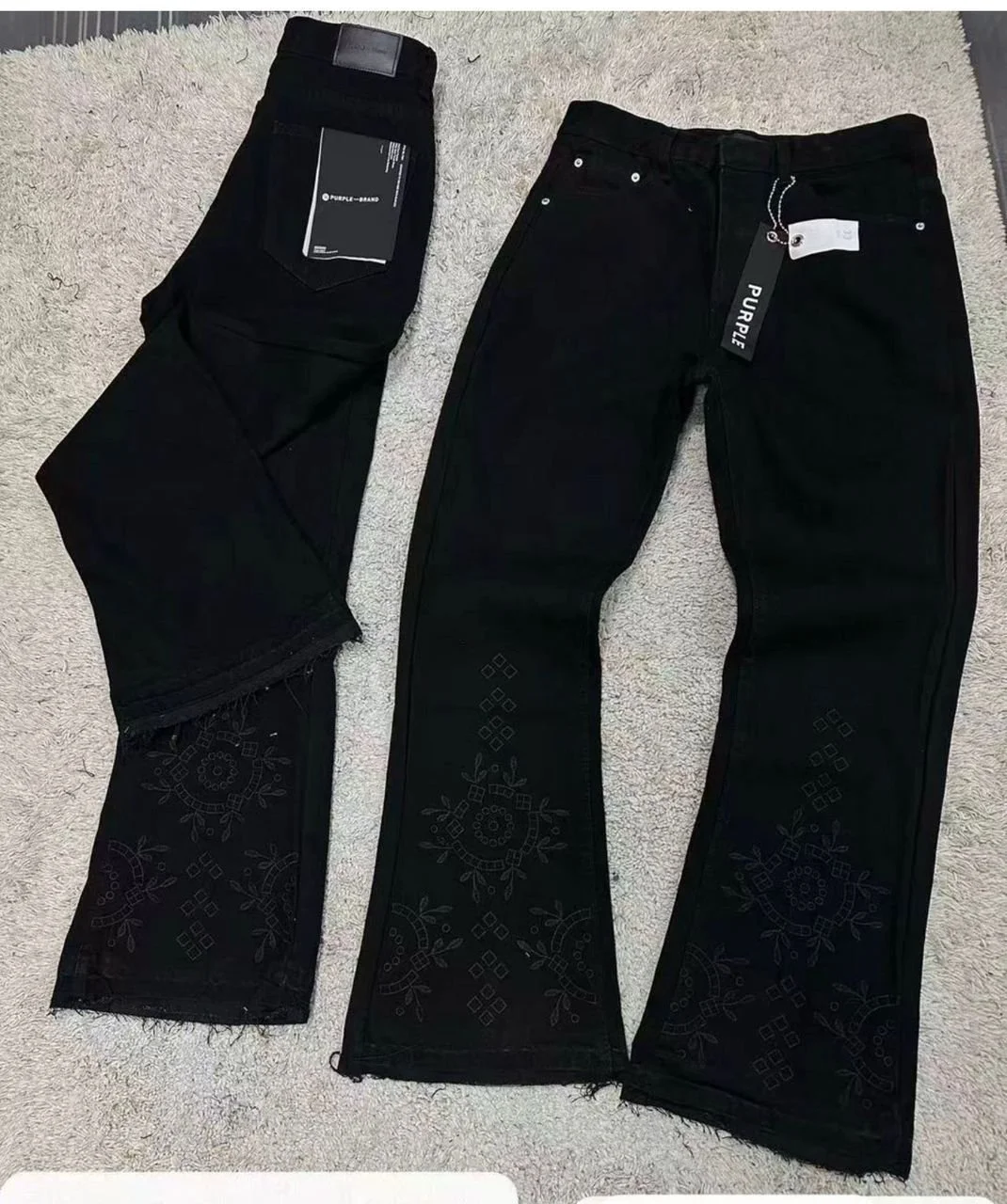 Two pairs of black pants, one with embroidery detail on the lower leg and the other with tags attached, lying on a light-colored carpet.