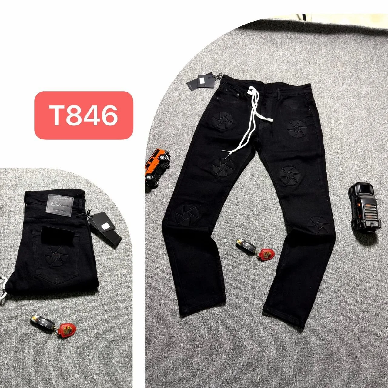 Pair of black jeans with patches laid out on a gray carpeted floor, with a white drawstring, key fobs, a toy car, and a folded pair of pants nearby.
