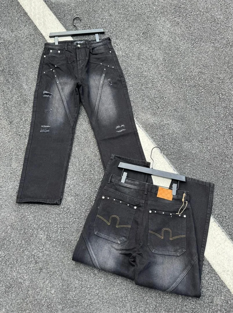 Pair of black distressed jeans with zipper details, one pair hanging on a hanger, and the other folded on the ground, on a paved surface.