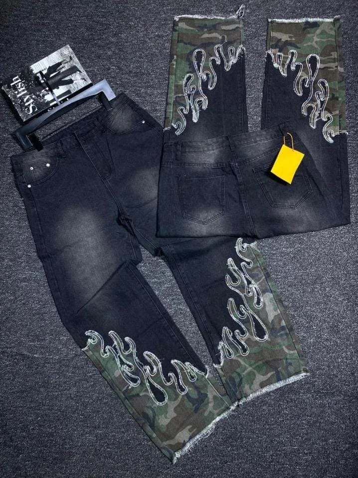 Pair of black jeans with flame embroidery and camouflage fabric accents, laid out on a gray carpet. An additional pair of camo accents with flame embroidery is also visible.