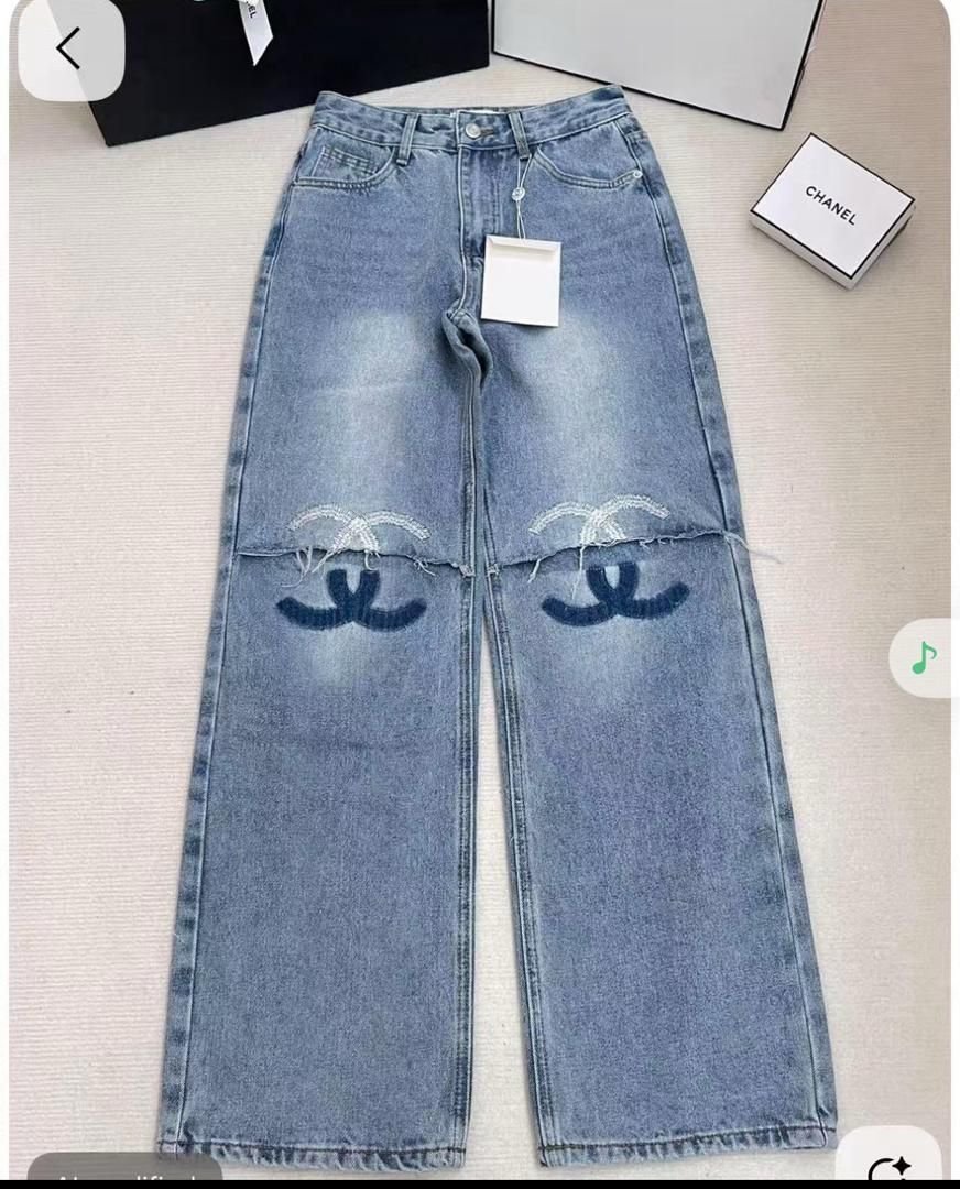 A pair of blue denim jeans with a distressed look, featuring a Chanel logo pattern near the knees, laid flat on a surface with Chanel boxes in the background.
