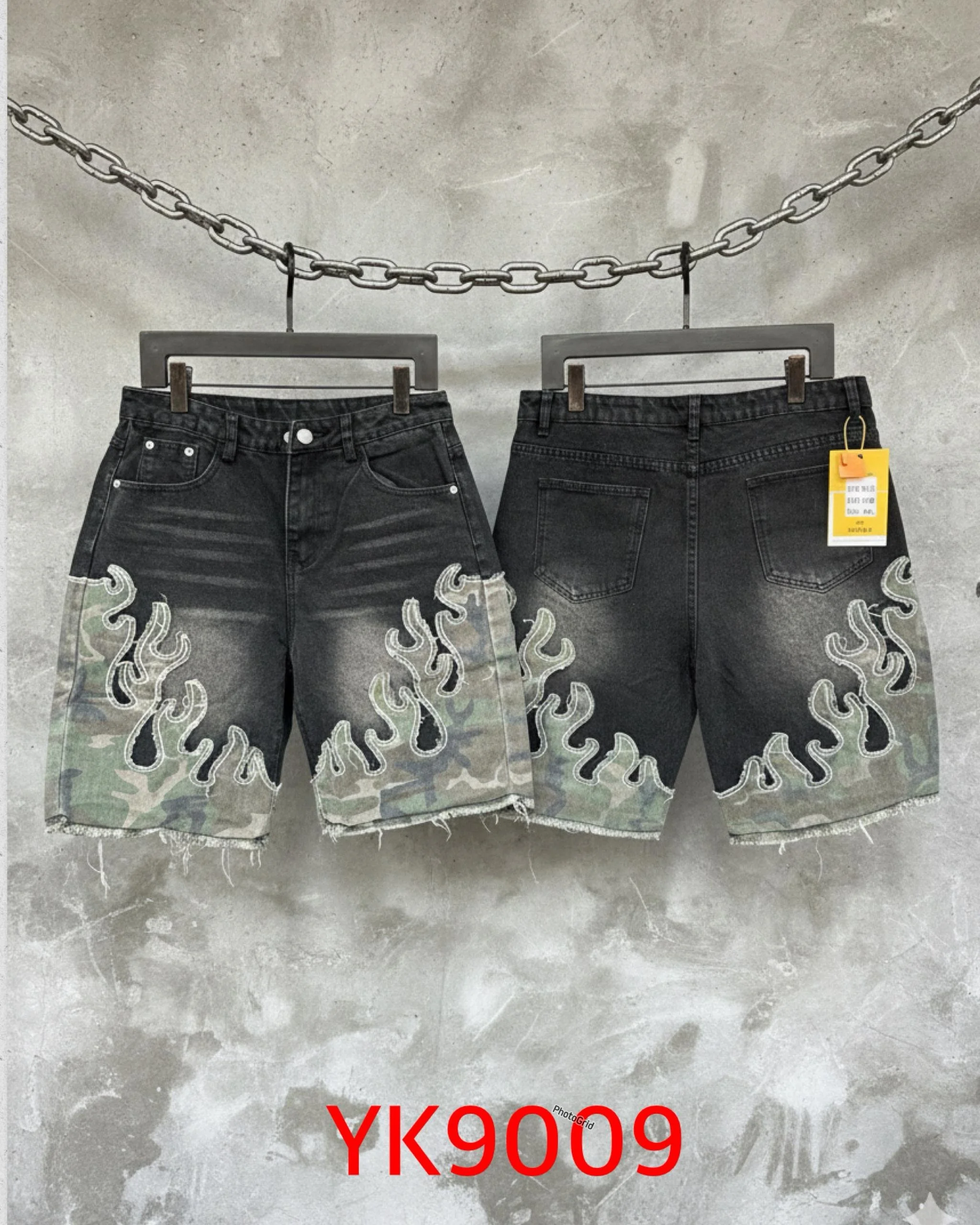 Pair of black denim shorts with flame and camouflage patterns, hanging on hangers in front of a concrete wall with a chain above.