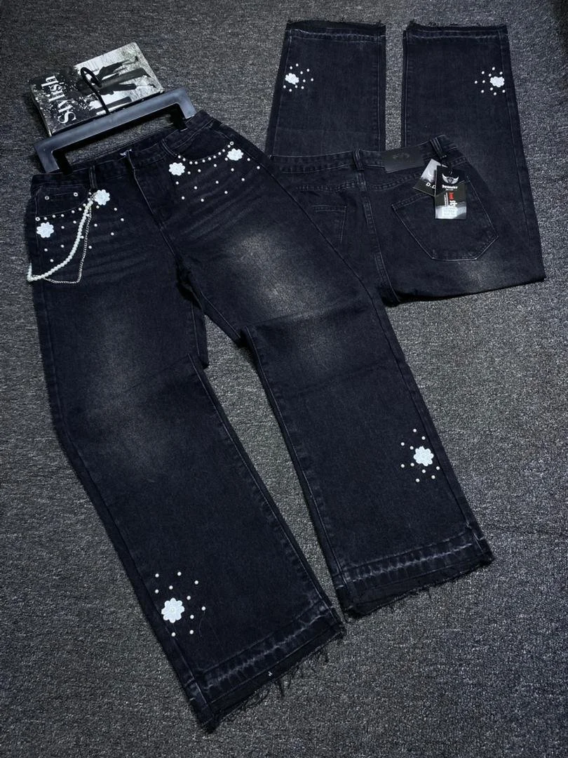 Pair of black jeans with white floral and dotted embellishments, laid flat on a gray carpet, one with a tag attached and a small accessory attached at the belt loop.