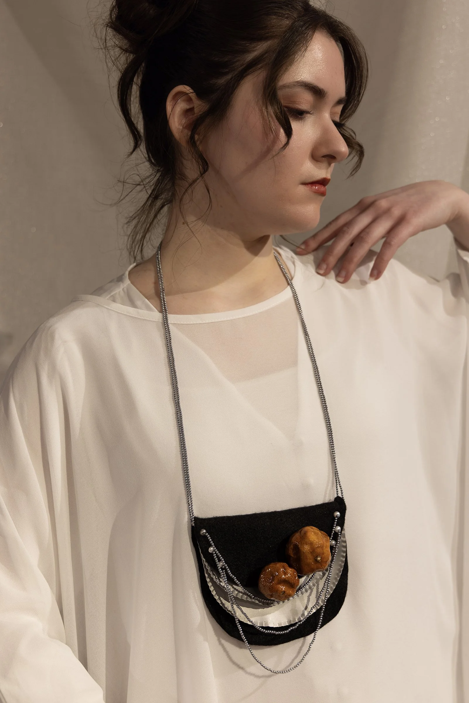 A woman with dark brown hair tied up, wearing a white top with sheer fabric in the front, and an unconventional necklace with objects such as oranges attached to a black fabric base, decorated with silver beads and chains.
