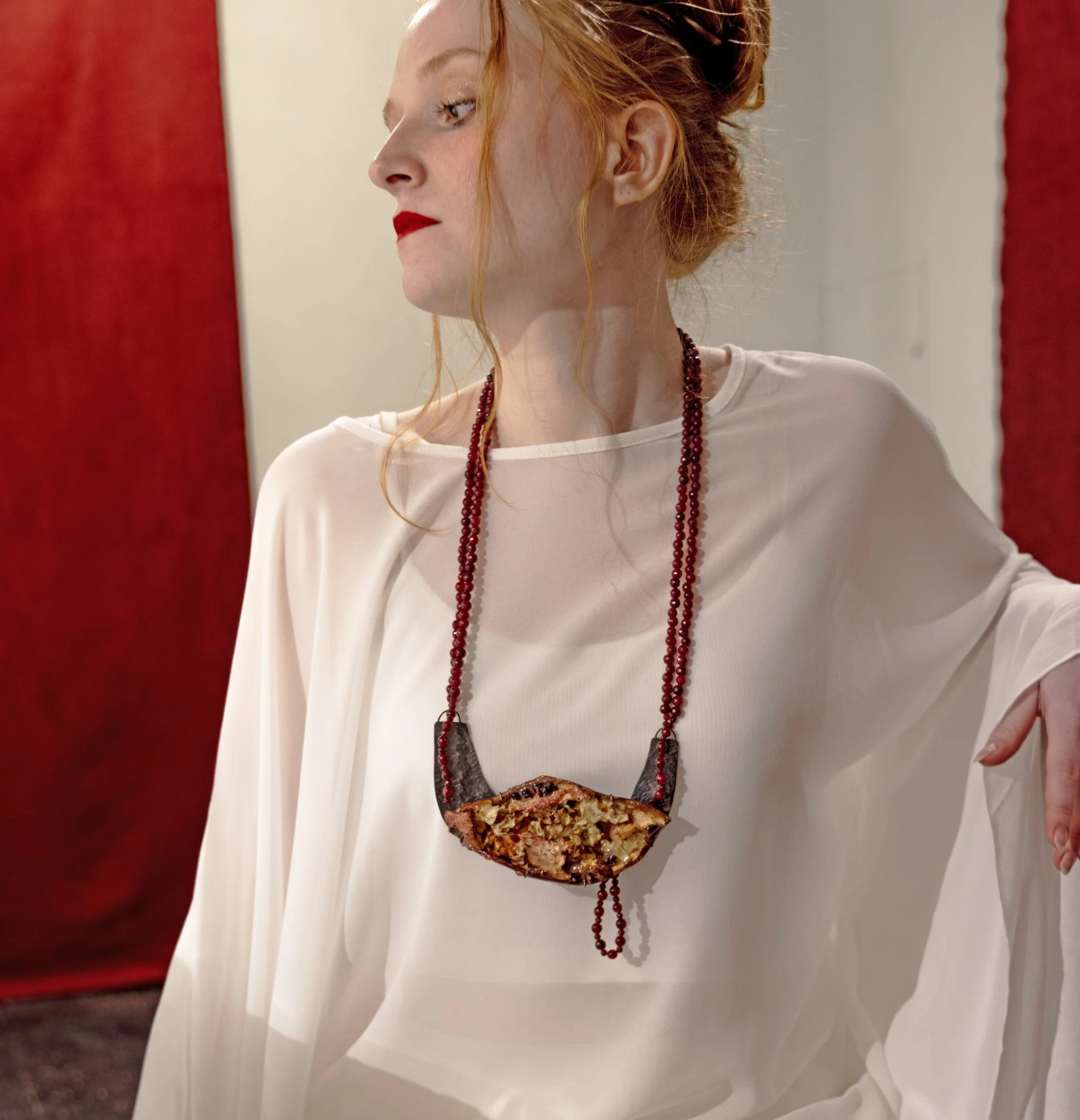 A woman with red hair wearing a white dress, red lipstick, and red beaded jewelry, standing against a blurred background of red and gray.