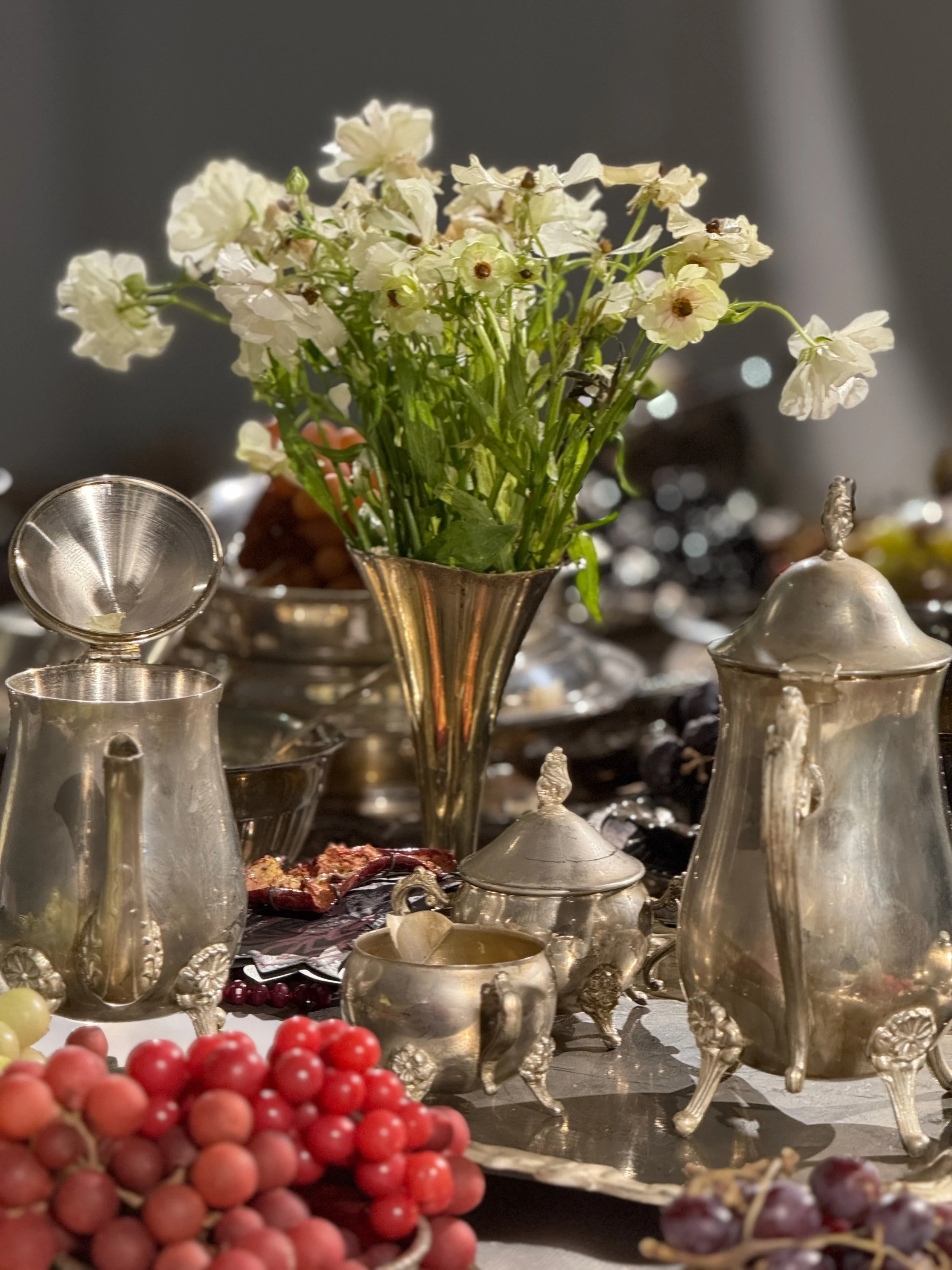 A floral arrangement of white flowers in a silver vase set on a table with silver and glass serving ware, with grapes in the foreground.