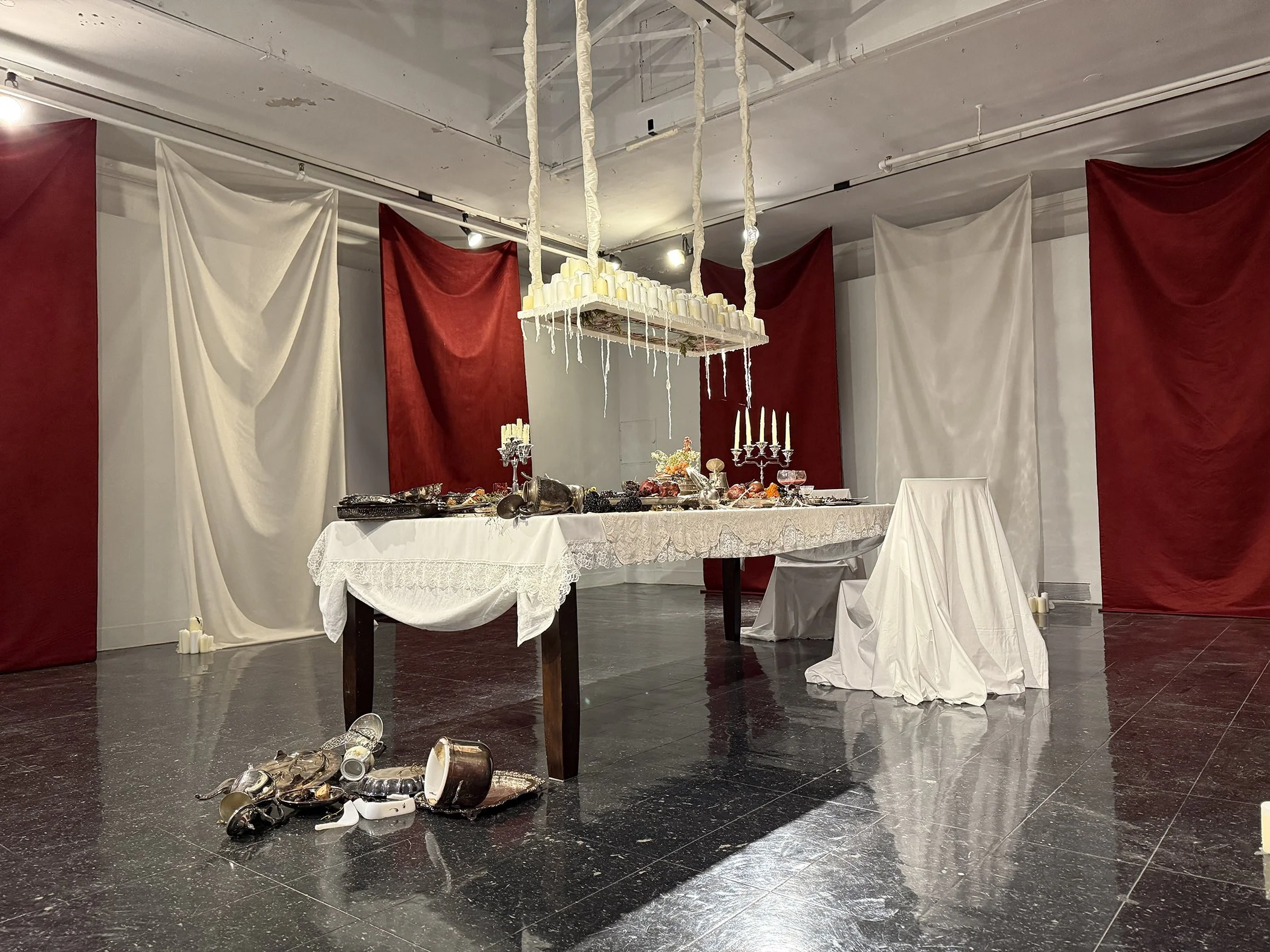 Disorderly table setup with spilled candles, shoes, cups, and scattered decorations in front of a backdrop of red and white draped fabric.