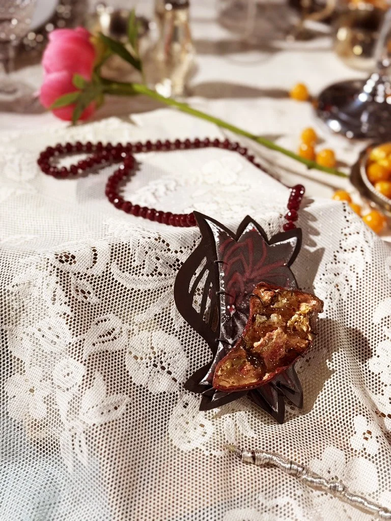 Decorative table setting with a lacy tablecloth, featuring a beaded enameled necklace, a pink flower, and dried pomnegranate.