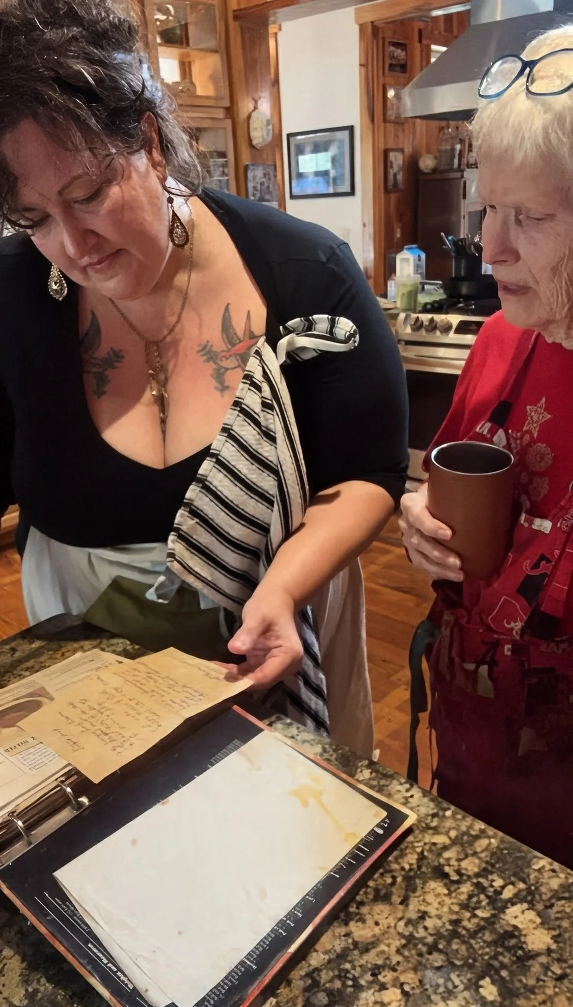 Two women, mother and daughter, looking at a cookbook on a kitchen counter, one woman holding a coffee mug.