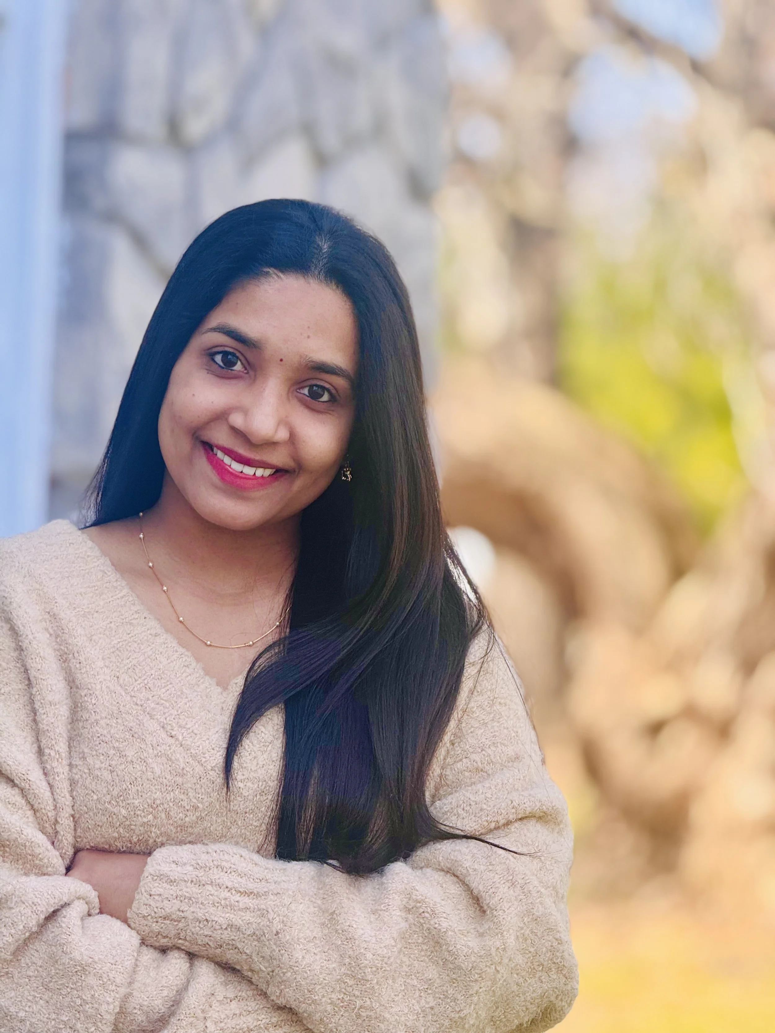A woman wearing a beige sweater with long black hair, smiling outdoors with blurred trees and stone wall in the background.