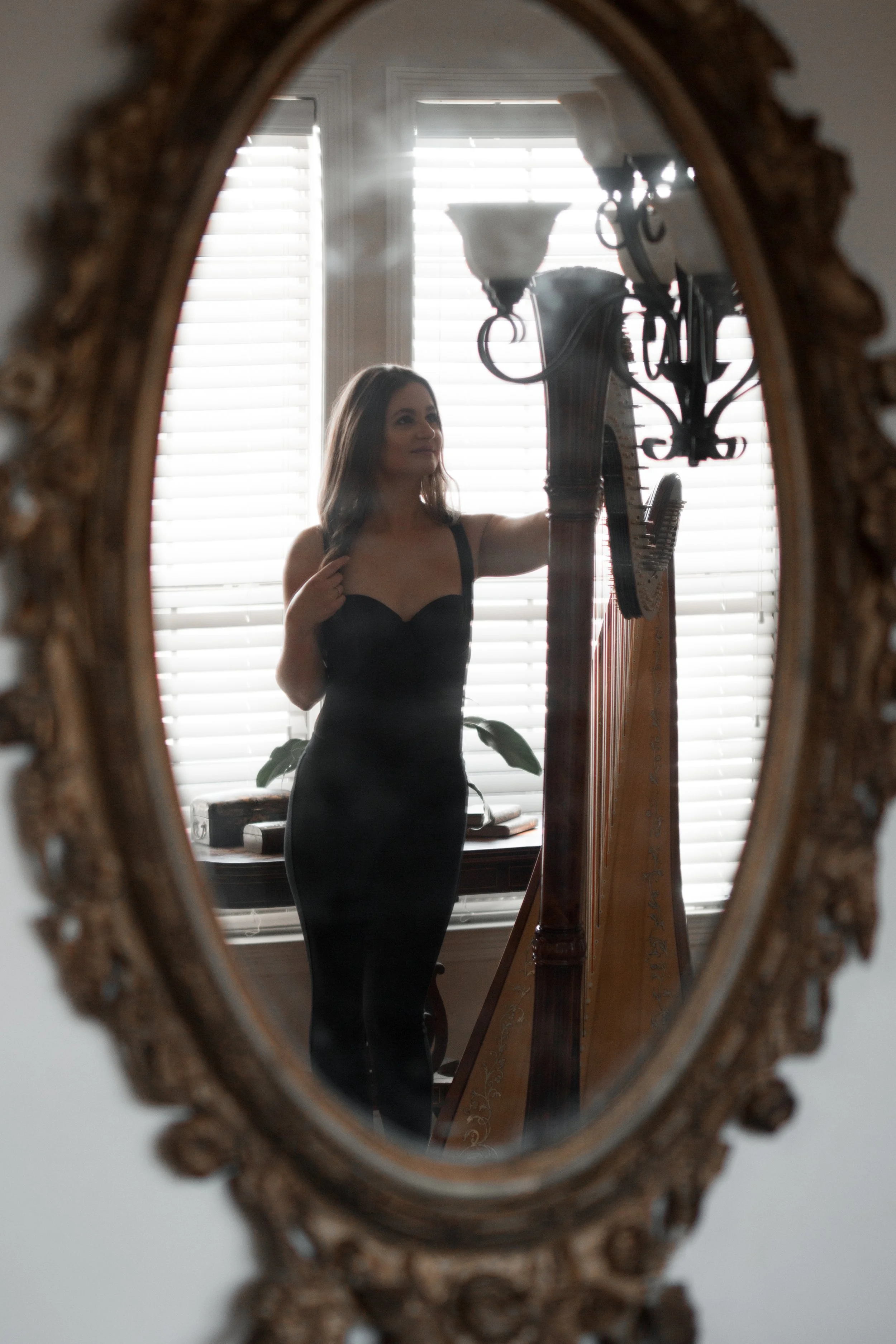 A woman in a black dress taking a selfie in an ornately framed mirror, with a harp and a window with blinds in the background.