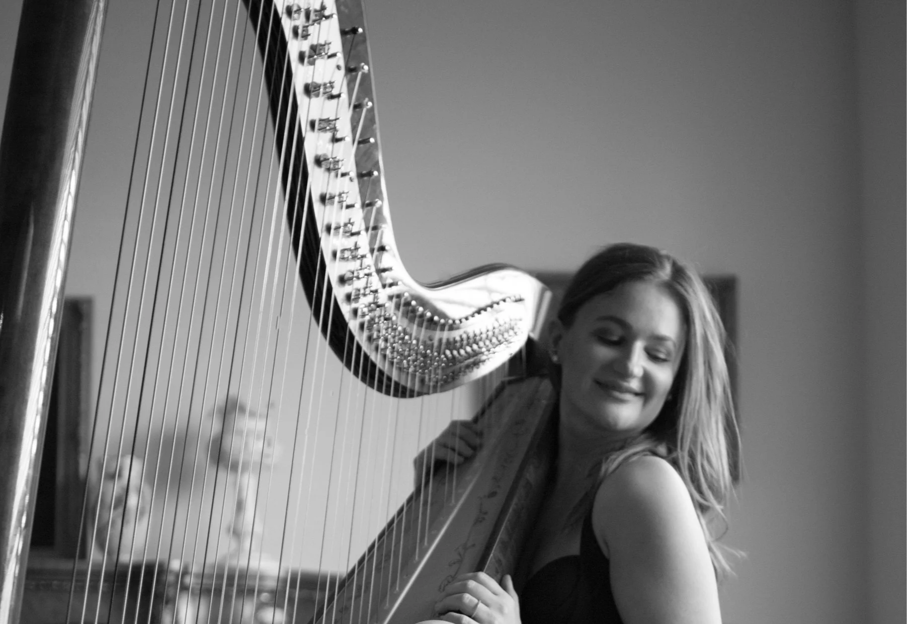 A black and white photo of a woman smiling while holding a harp, with the strings and part of the instrument visible.