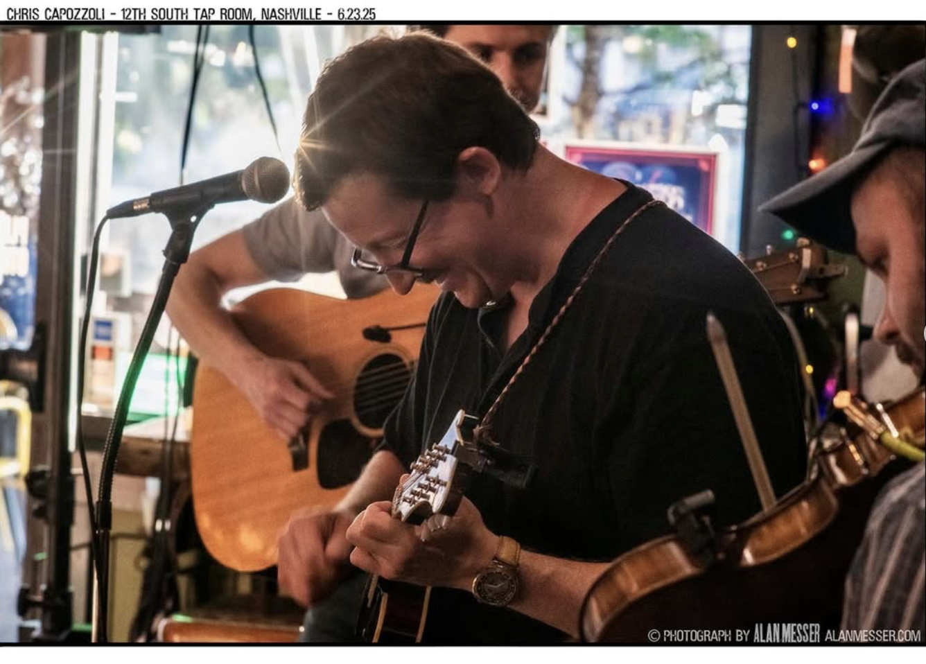 Three musicians performing indoors: one playing acoustic guitar, another on electric guitar, and a third on electric violin, with colorful lights in the background.
