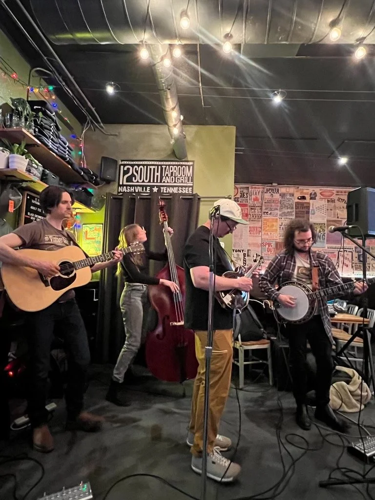 A four-member band performing live in a small, cozy music venue. The band includes a guitarist, bassist, banjo player, and another guitarist. The venue has colorful posters on the wall, string lights overhead, and a sign that reads '12 South Taproom and Grill, Nashville, Tennessee.'