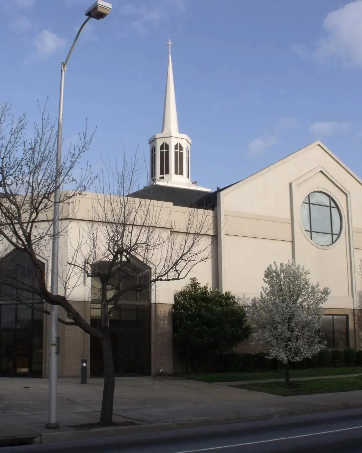 A church building with a tall steeple topped with a cross, a round window, leafless trees, a flowering tree, and a streetlight against a blue sky.