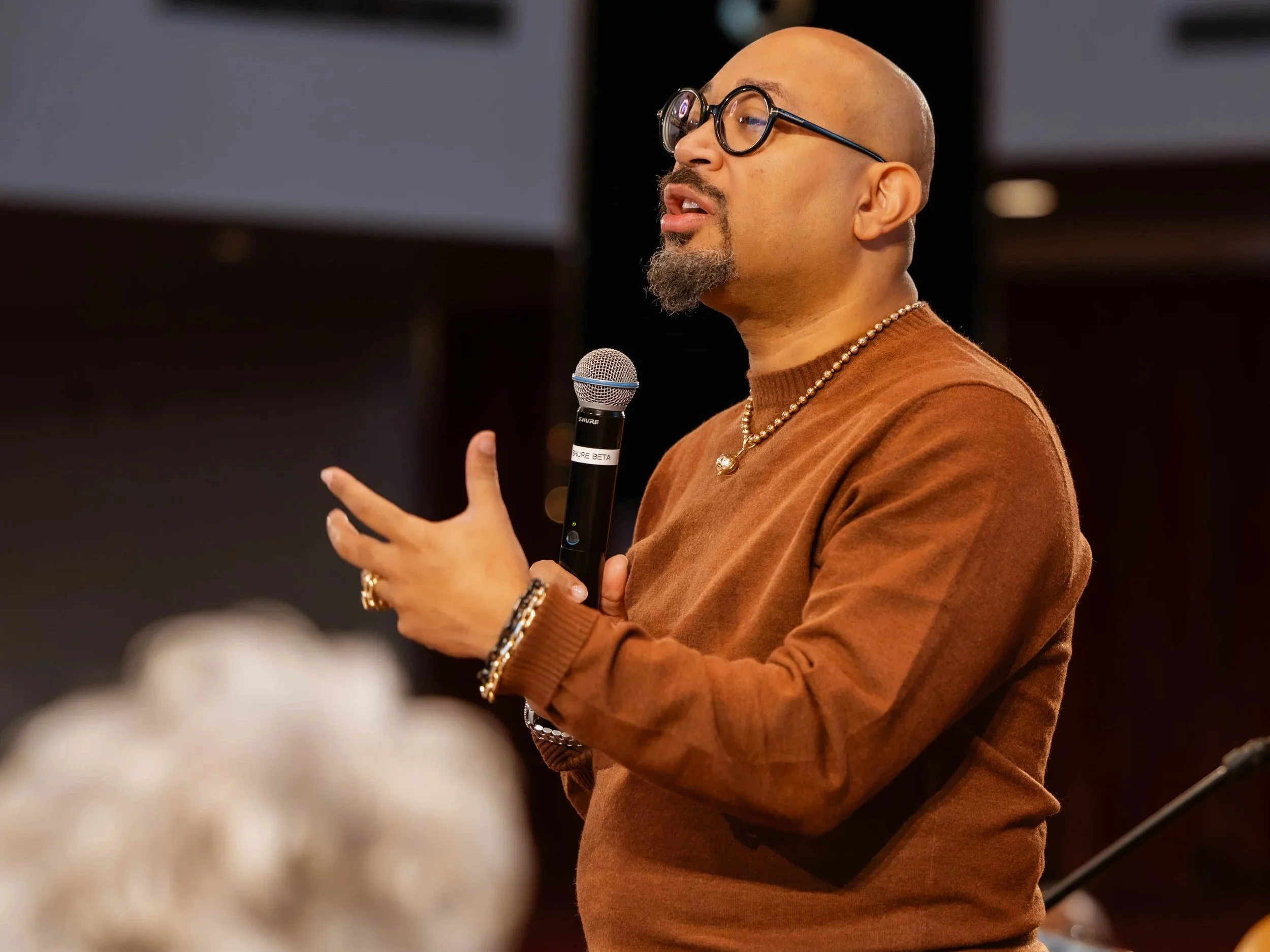 Man speaking into a microphone during a presentation or speech, wearing glasses, a brown sweater, and jewelry, with a blurred audience in the foreground.