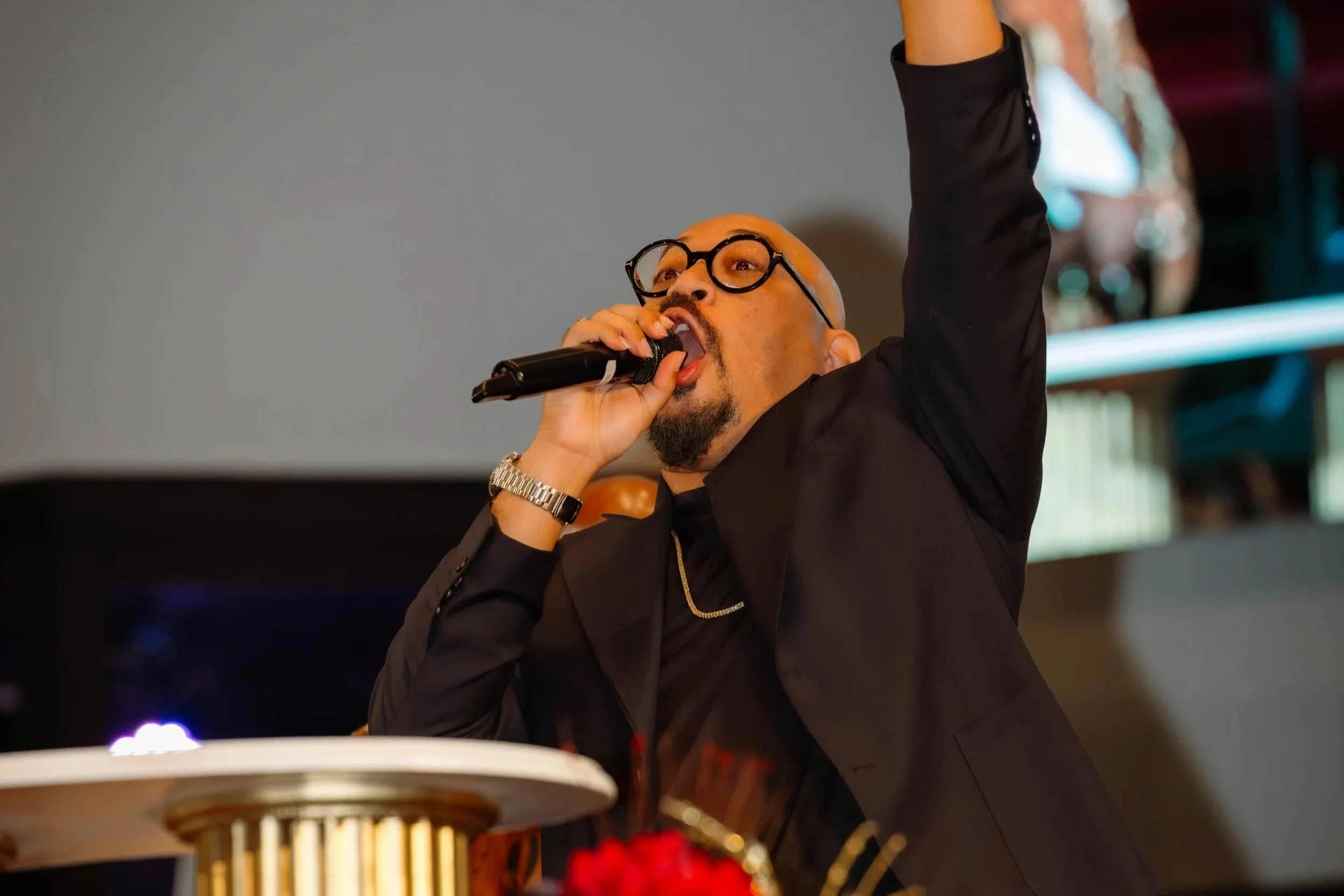 A man wearing glasses and a black jacket, holding a microphone and singing or speaking. Part of a birthday cake with candles is visible in the foreground.
