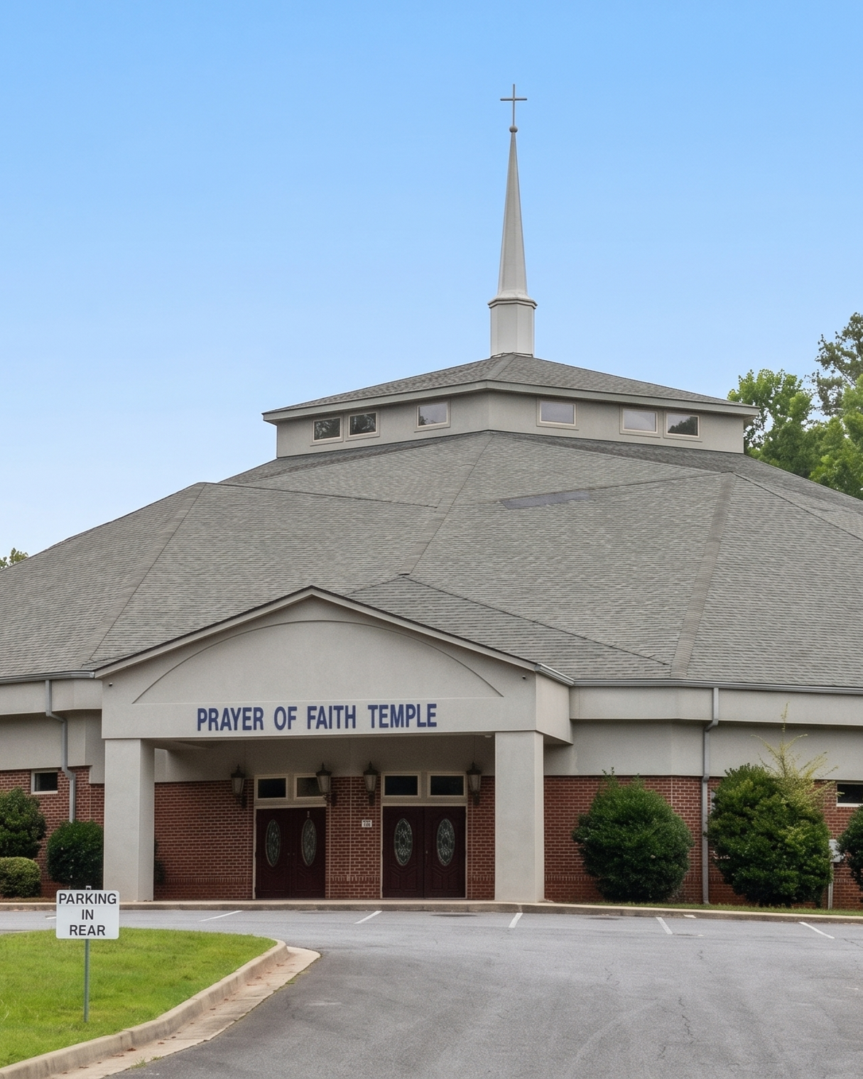 Front view of the Prayer of Faith Temple with a brick and white exterior, a sign above the entrance, and a steeple with a cross on top, against a blue sky.