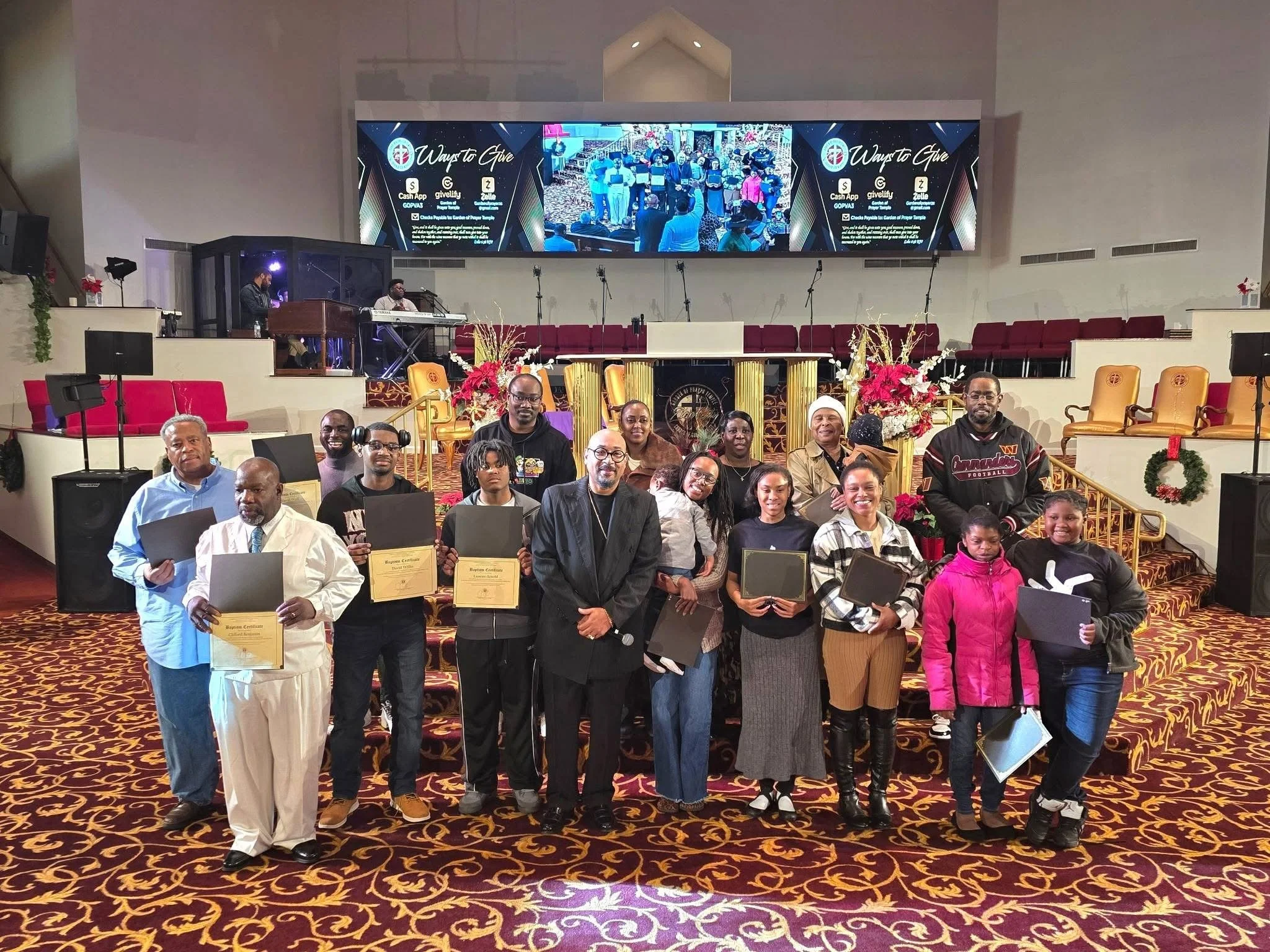 A group of people gathered in a church, holding certificates, with a large digital screen displaying worship service information and a photo of the congregation in the background. The church is decorated with flowers and Christmas wreaths.