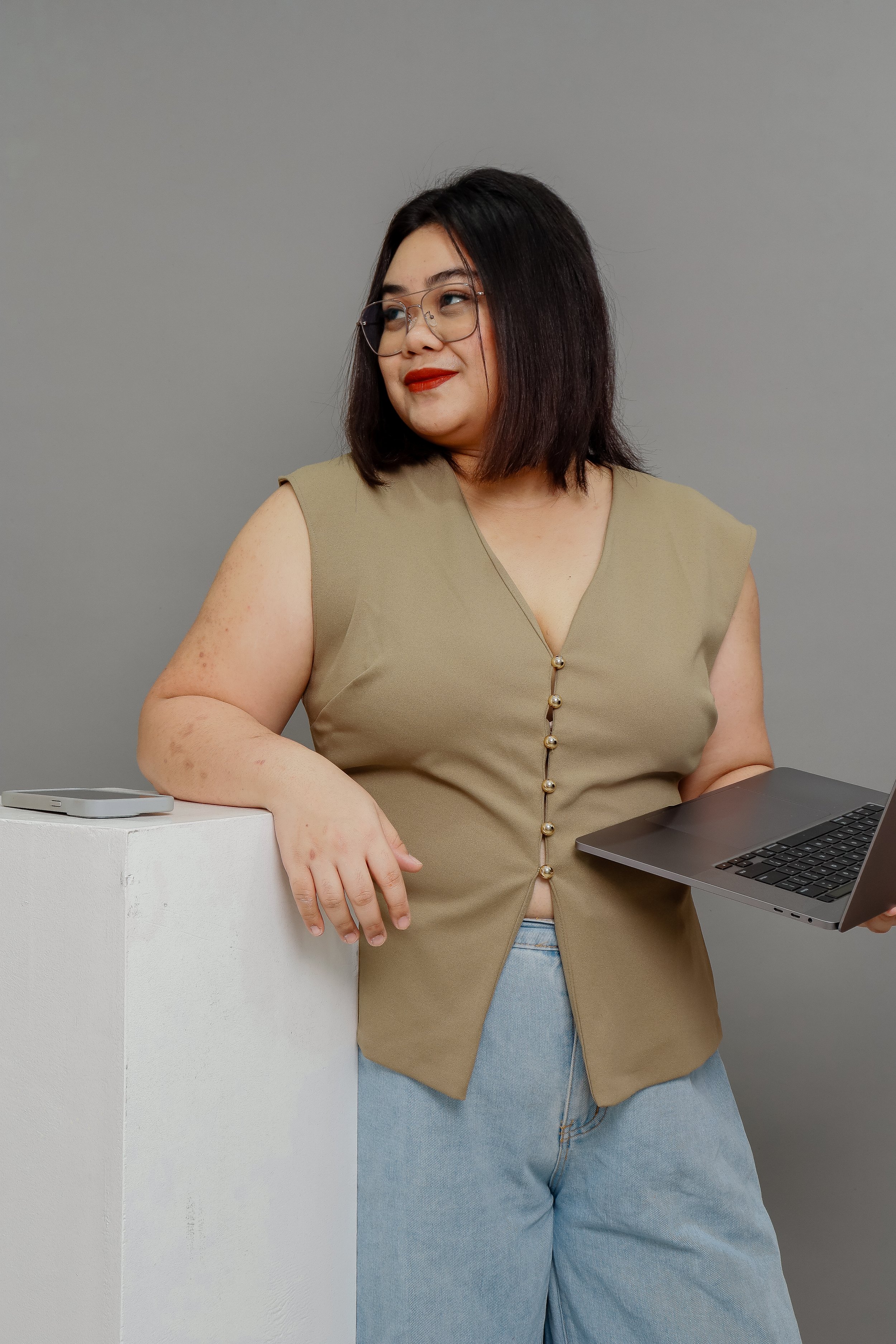 A woman with shoulder-length dark hair, glasses, and red lipstick, wearing a sleeveless beige top with pearl buttons and light blue jeans, standing next to a white pedestal with a smartphone on top, holding a laptop in her right hand, against a plain gray background.