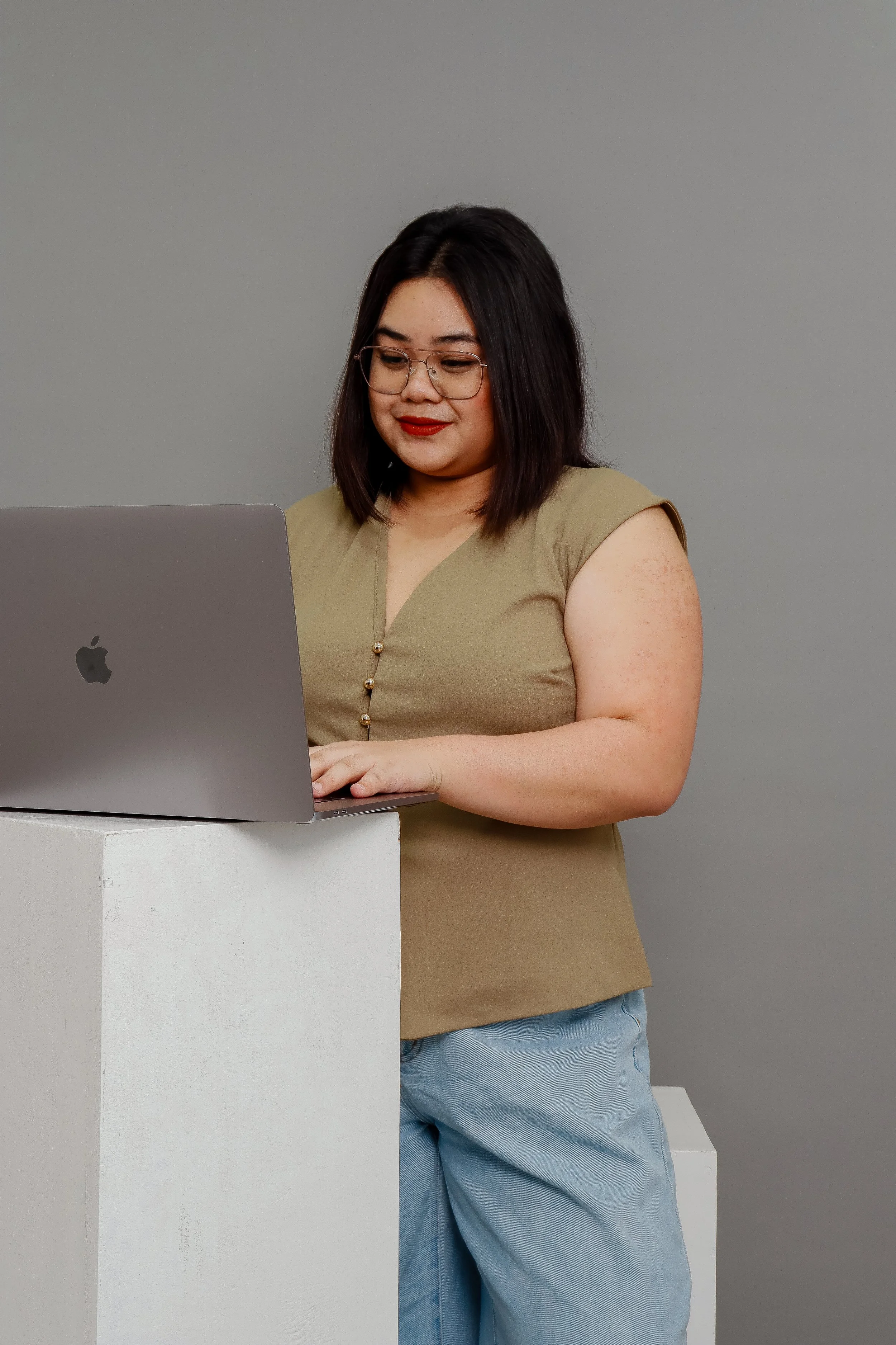 Woman with black hair and glasses wearing a beige top and blue jeans, standing behind a white podium and working on a silver MacBook laptop, against a plain gray background.