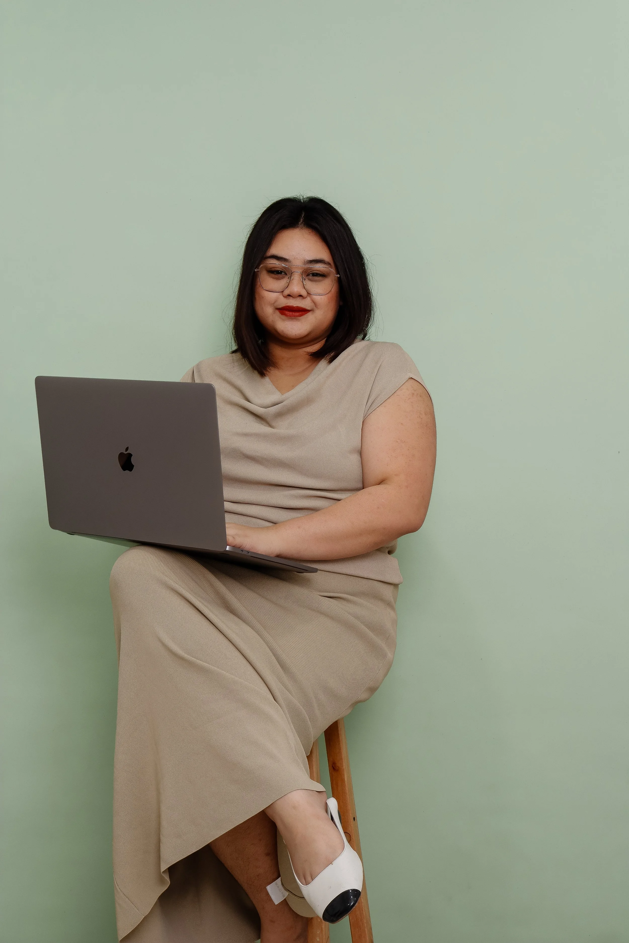 A woman with dark hair, glasses, and red lipstick sitting on a wooden stool, looking at a silver MacBook, against a light green wall, in a beige dress and beige shoes.