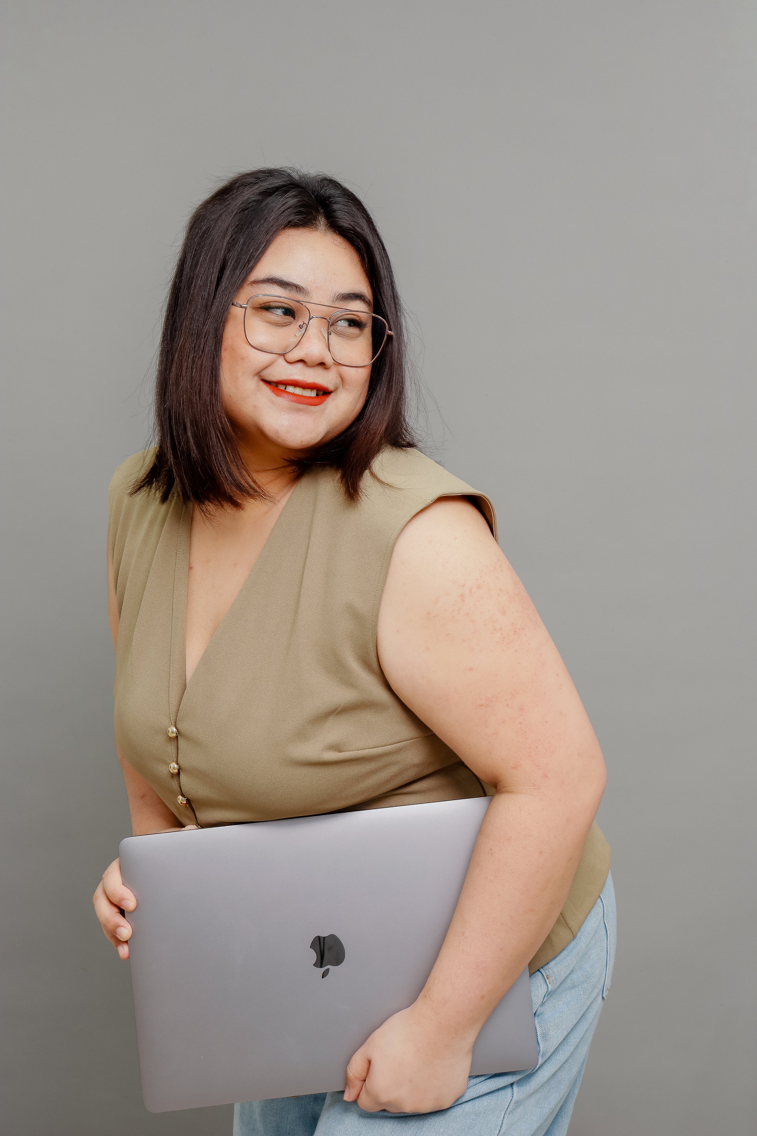 A woman with shoulder-length dark hair, glasses, and red lipstick, looking to her right while holding a closed silver MacBook in front of her. She is wearing a beige sleeveless top and light blue jeans, standing against a plain gray background.