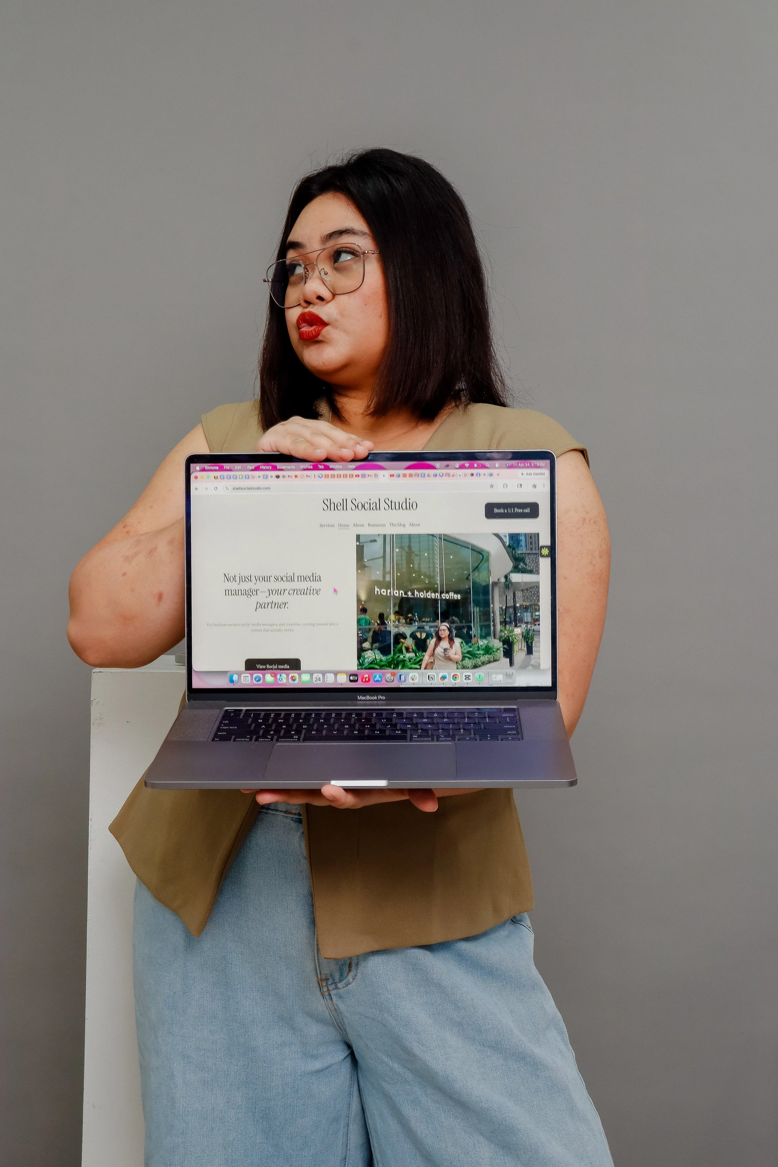 A woman with dark hair, glasses, and red lipstick holding a laptop in front of her. The laptop shows a website called 'Shell Social Studio' featuring a coffee shop. She is wearing a tan sleeveless top and light blue jeans, standing against a plain gray background.
