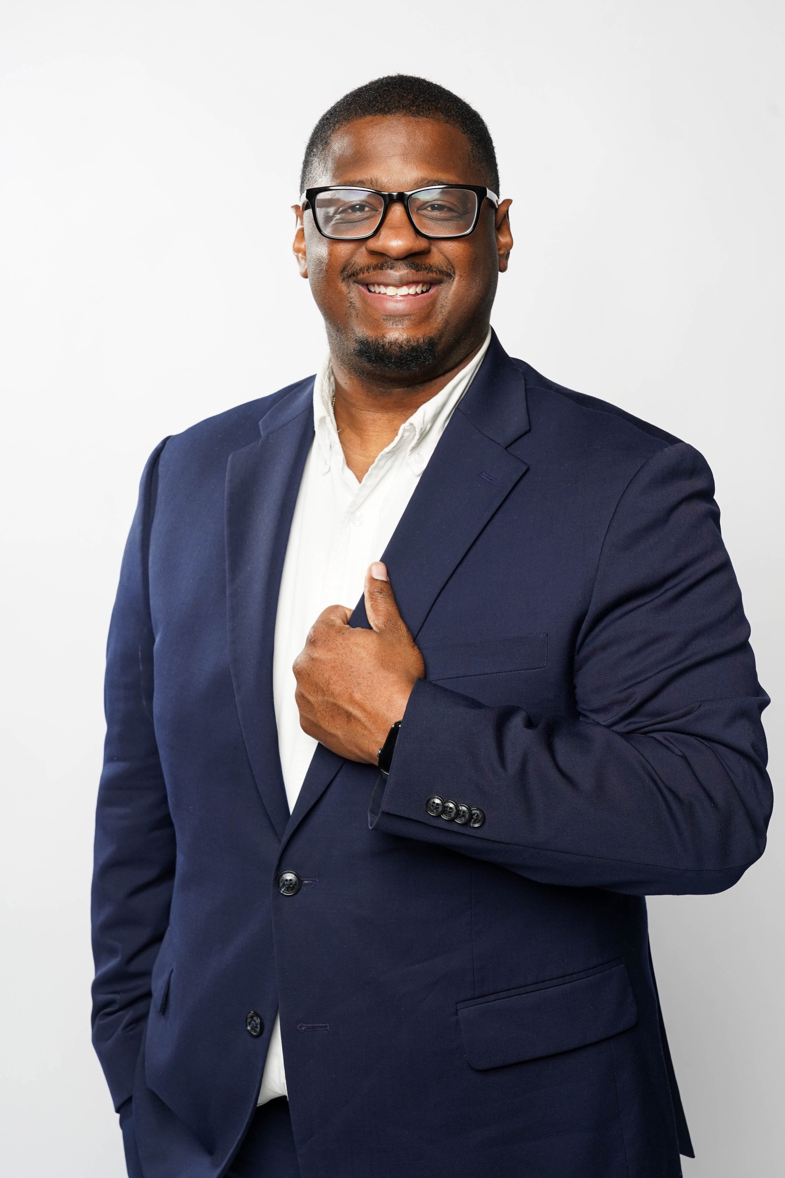 Portrait of a smiling man wearing glasses, a navy blue suit, and a white shirt, standing against a plain white background. Keenan R. Lowery.