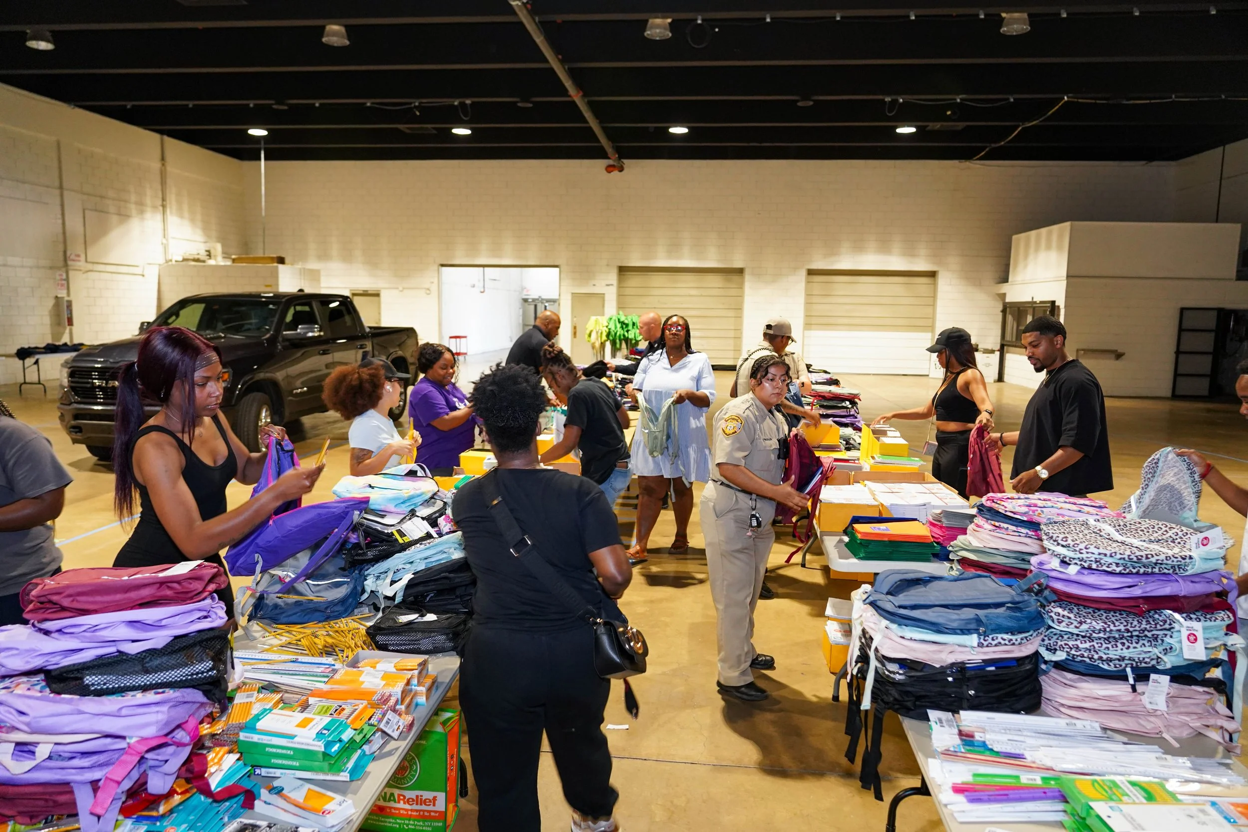 People shopping for school supplies at a donation event, with tables filled with notebooks, folders, and backpacks in a large indoor space.