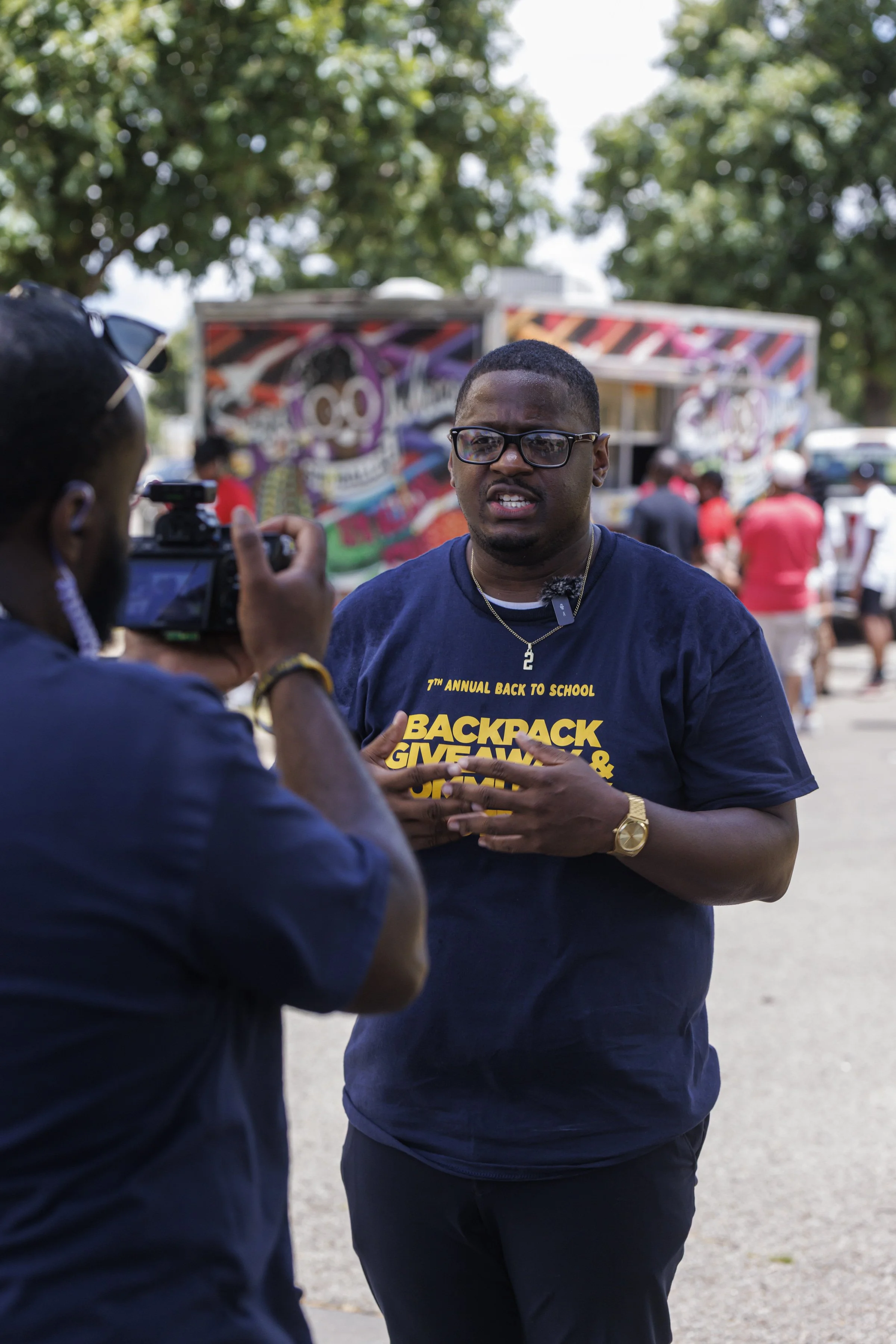 A man wearing glasses, a navy T-shirt, and a gold watch being interviewed by a person holding a camera. The T-shirt reads "7th Annual Back to School Backpack Giveaway." Keenan R. Lowery and Kinnawa Kaitibi are capture by MadebyGlyde