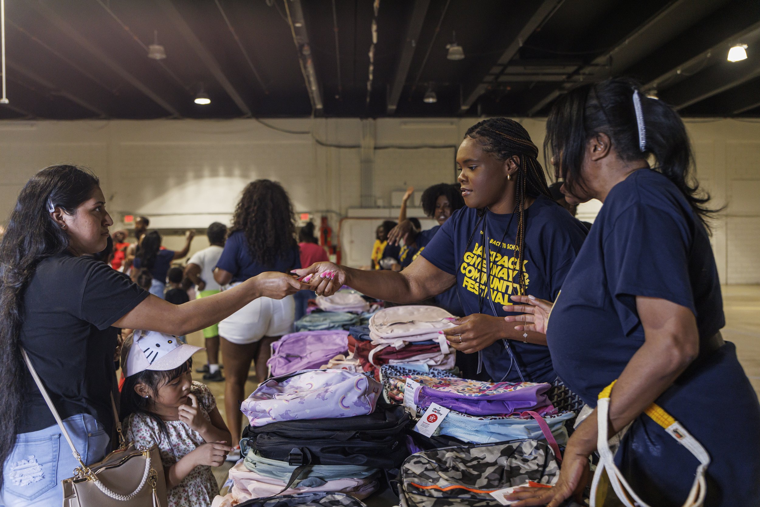 People at a tables with backpacks and clothing during a community event or fair inside a gymnasium, interacting and exchanging items.