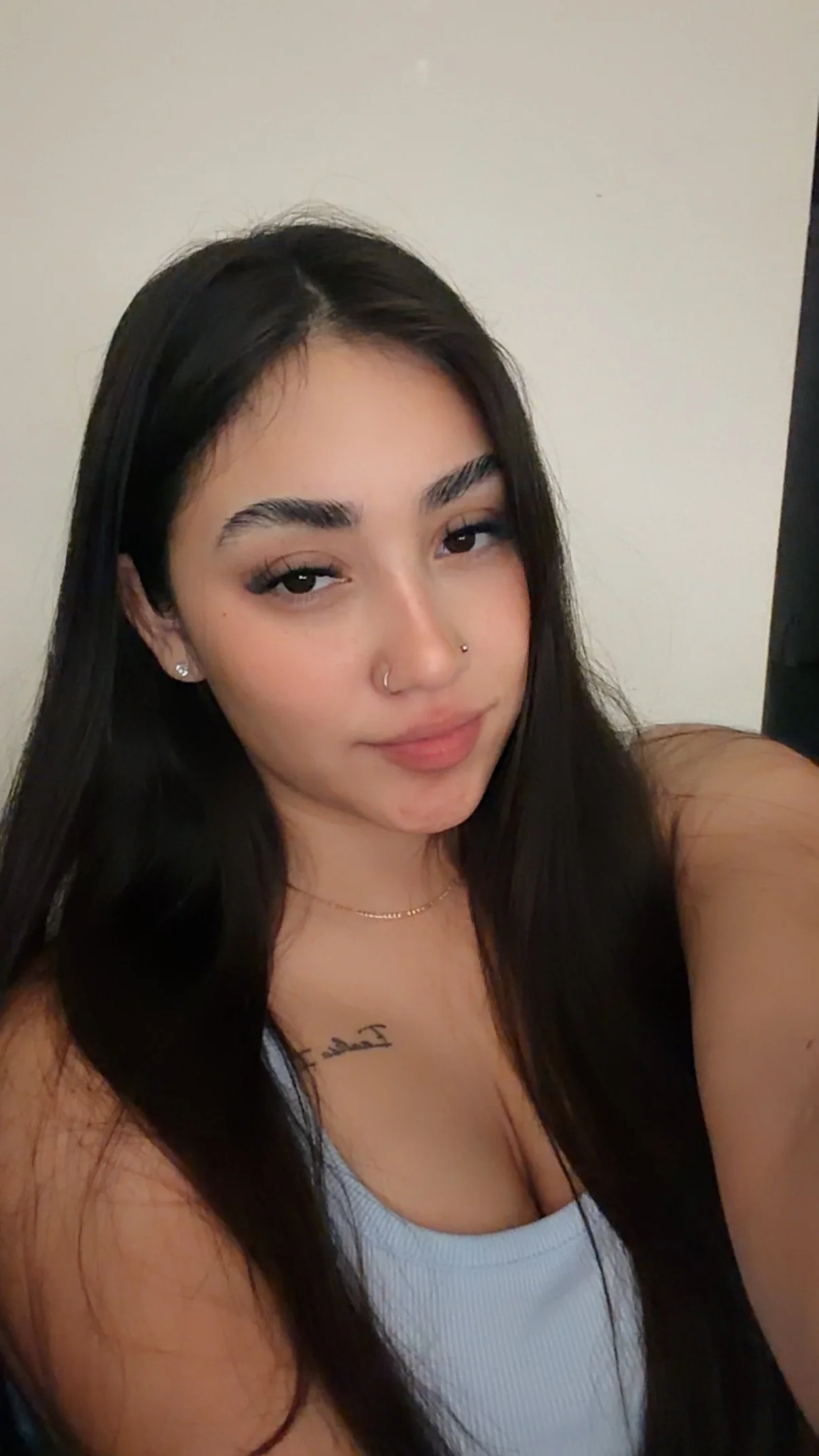 A young woman with long black hair taking a selfie in front of a plain wall, wearing a white top, with a tattoo visible on her chest.