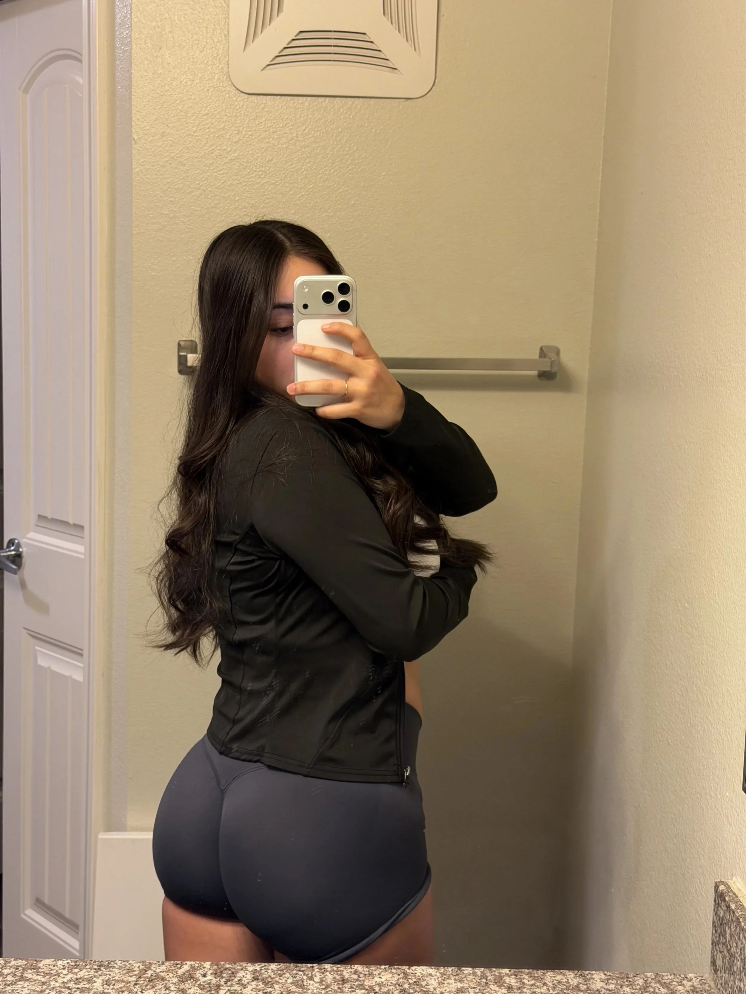 A young woman taking a mirror selfie in a bathroom. She has long dark hair, wearing a black jacket and dark gray shorts, and is holding her phone in front of her face.