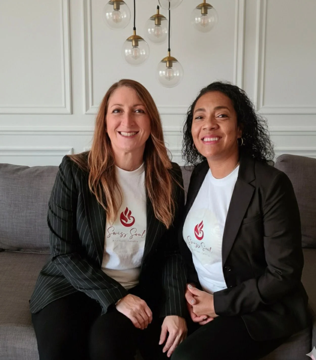 Zwei Frauen sitzen auf einem grauen Sofa, tragen weiße T-Shirts mit rotem Logo und schwarzen Blazer, lächeln und posieren für die Kamera, im Hintergrund weiße Wand mit Wandpaneelen und einer modernen Deckenleuchte.
