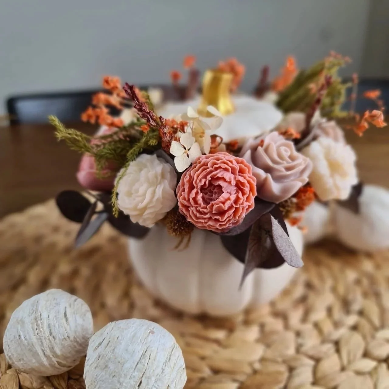 Blumengesteck mit Rosen, Eukalyptus und anderen trockenen Blumen in einer weißen Vase, auf einem Holztisch mit zwei weißen dekorativen Ball-Objekten.